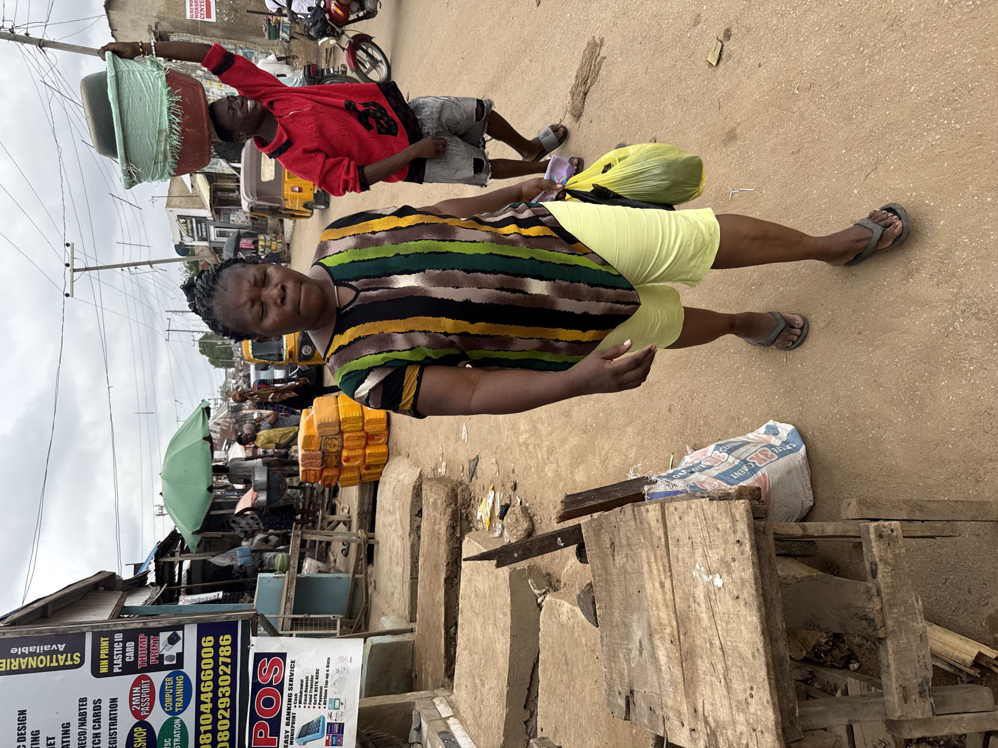 The image depicts a street scene in what appears to be a commercial or market area of Abuja, Nigeria. The street is unpaved and the scene is bustling with commercial activity.
In the foreground, a woman is walking toward the viewer, carrying a yellow bag. Behind her and slightly to the left, a man carries a stack of pots on his head.
To the left of the frame are several market stalls and shops, identifiable by their signage. One sign advertises stationery and banking services. Adjacent to the stalls are several plastic containers and an green umbrella. A set of low, stepped structures constructed of what appears to be dried clay lead up to the stalls.
The weather appears overcast, and the scene is filled with details indicative of a vibrant, active market environment.