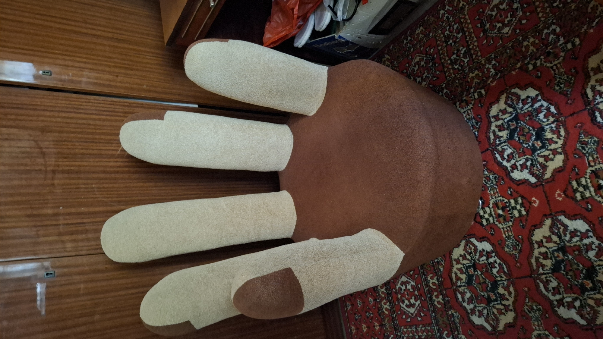 An unusual armchair, designed in the shape of a hand with outstretched fingers, sits on a red oriental rug in a room with wooden cabinetry. The armchair is upholstered in brown fabric for the palm and thumb, with beige fabric for the fingers. The rug features a complex pattern of red, cream, and black. The wooden cabinetry appears to be built-in, with visible doors and handles. To the right of the armchair, a portion of a wooden desk or cabinet is visible, along with some stacked items, including a red plastic bag and what appears to be white footwear. The scene suggests an indoor residential setting. The exact location in Shymkent, Kazakhstan cannot be determined from the image alone.
