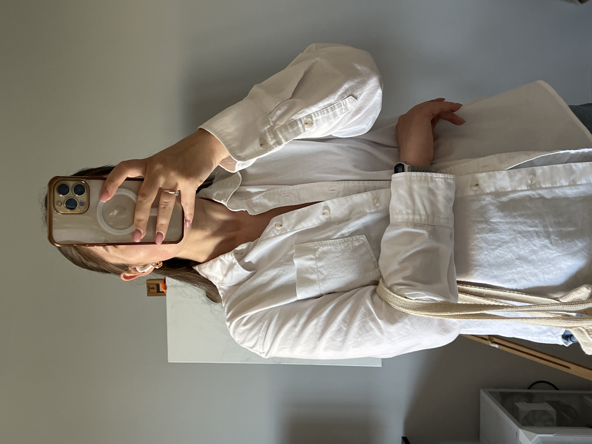 A person with fair skin and brown hair is taking a selfie using a gold-colored smartphone. The person is wearing a white button-down shirt with long sleeves, a ring on one finger, earrings, and a watch on their wrist. The background consists of a grey wall, a white cabinet, and glimpses of other objects. There is no discernible evidence in the image to indicate the location is in Ternopil, Ukraine.