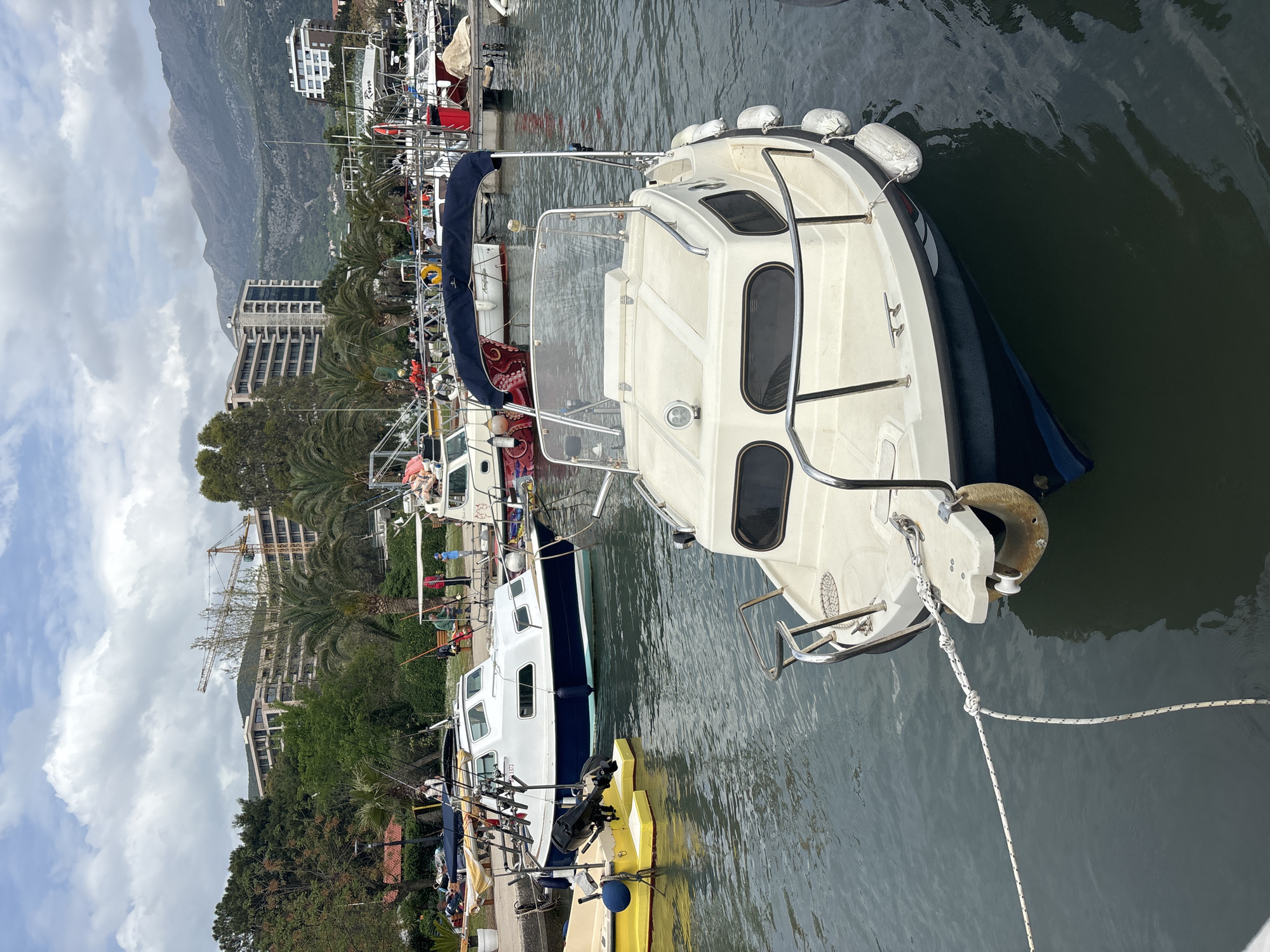 The image depicts a marina in Budva, Montenegro. The scene includes several boats docked in the water. The primary boat, closer to the viewer, is a white motorboat with a blue accent and a cabin. Along the dock, to the left of the white boat, other boats are visible, including a yellow kayak docked near a boat with multiple fishing rods. The water appears calm, reflecting the surroundings. In the background, buildings, palm trees, and a crane are visible, indicating an urban area with construction. Mountains are also present in the background, suggesting a coastal city. The sky is partially cloudy.