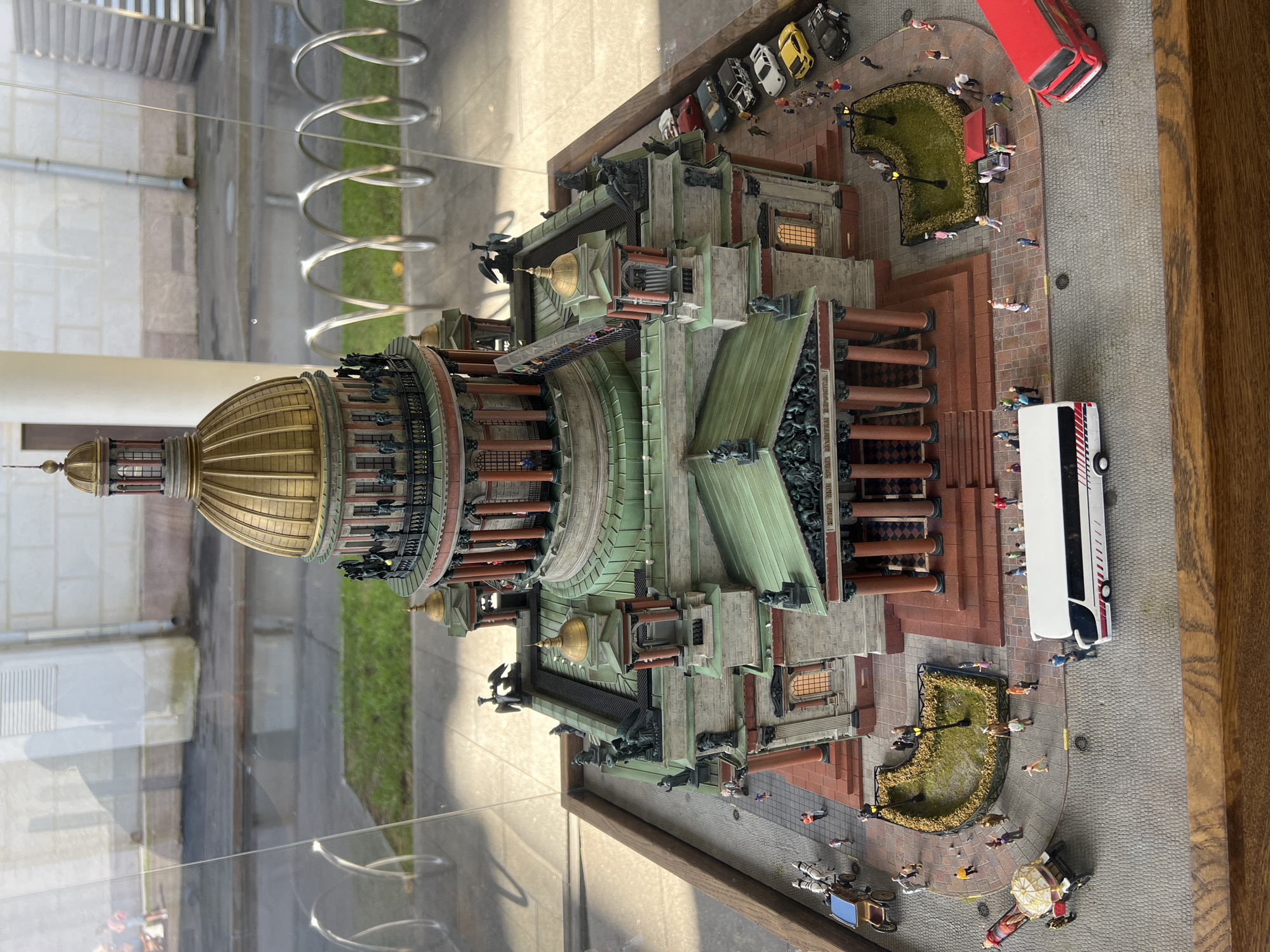 The image shows a detailed miniature model of Saint Isaac's Cathedral, a prominent landmark in St. Petersburg, Russia. The model is displayed inside a glass enclosure, with reflections visible on the glass surface, suggesting it is located in an indoor exhibition space or museum. The model features intricate architectural details of the cathedral, including its golden dome, classical columns, and decorative statues. Surrounding the cathedral model is a diorama depicting a street scene with miniature figures of people, vehicles such as a bus, a red double-decker bus, cars, and a horse-drawn carriage, and landscaping elements like trees and flower beds. The overall scene is designed to represent the area around the actual cathedral, with cobblestone streets and sidewalks. The scene appears to be a static display intended to showcase the architectural grandeur of Saint Isaac's Cathedral and its surrounding urban environment. The background of the image shows out-of-focus elements that could be part of the museum's interior or the view outside the enclosure. It's important to note that Saint Isaac's Cathedral is located in St. Petersburg, not Moscow.