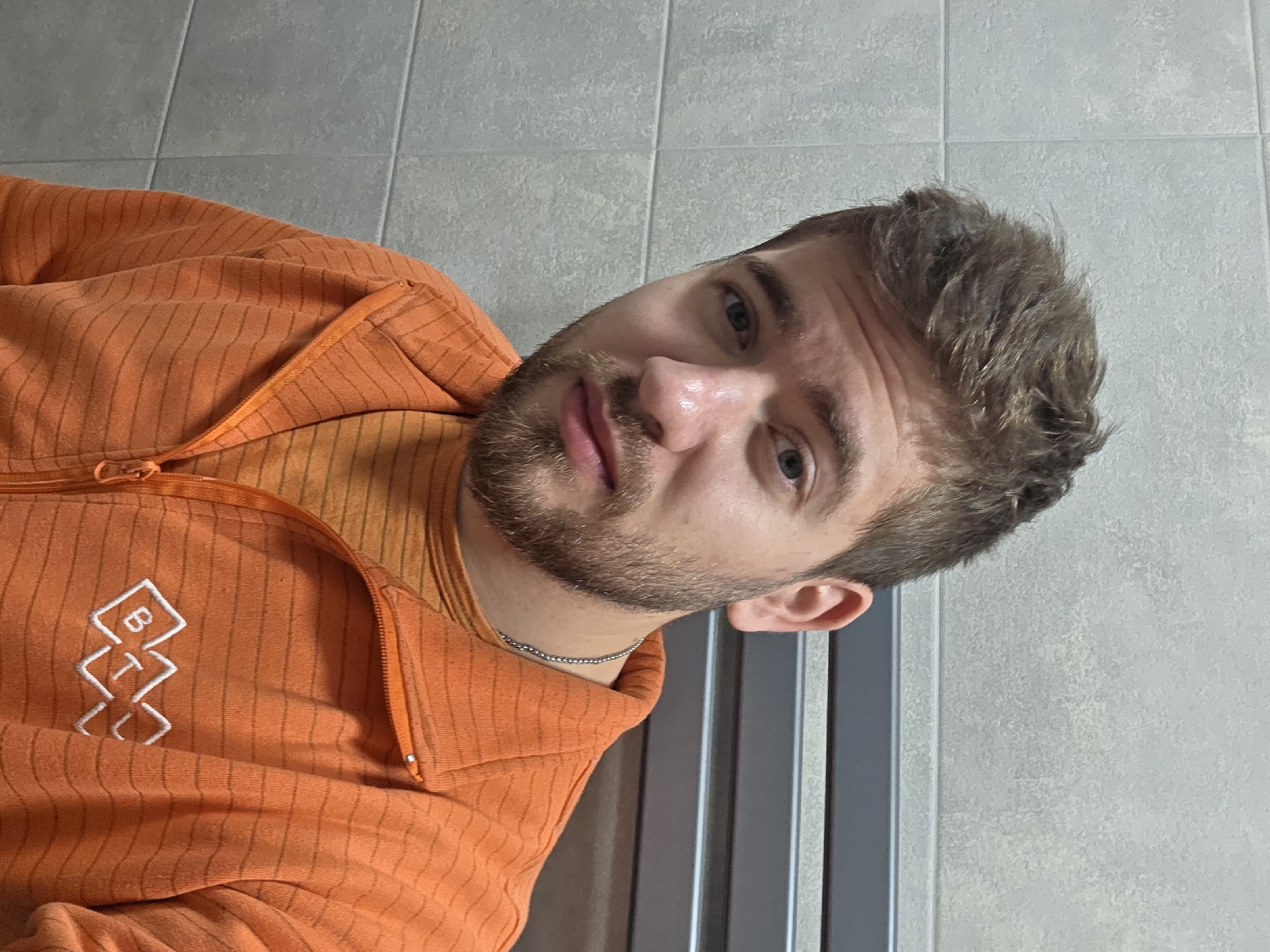 A bearded man in an orange, striped, collared, and zippered shirt with a white logo on the left breast is depicted indoors, likely a bathroom. The man's face is angled slightly upward and to the left. He has short, light brown hair and stubble. Behind him is a light gray tiled wall and a dark gray vertical radiator. There are no immediate indicators of Yagodovo, Bulgaria.