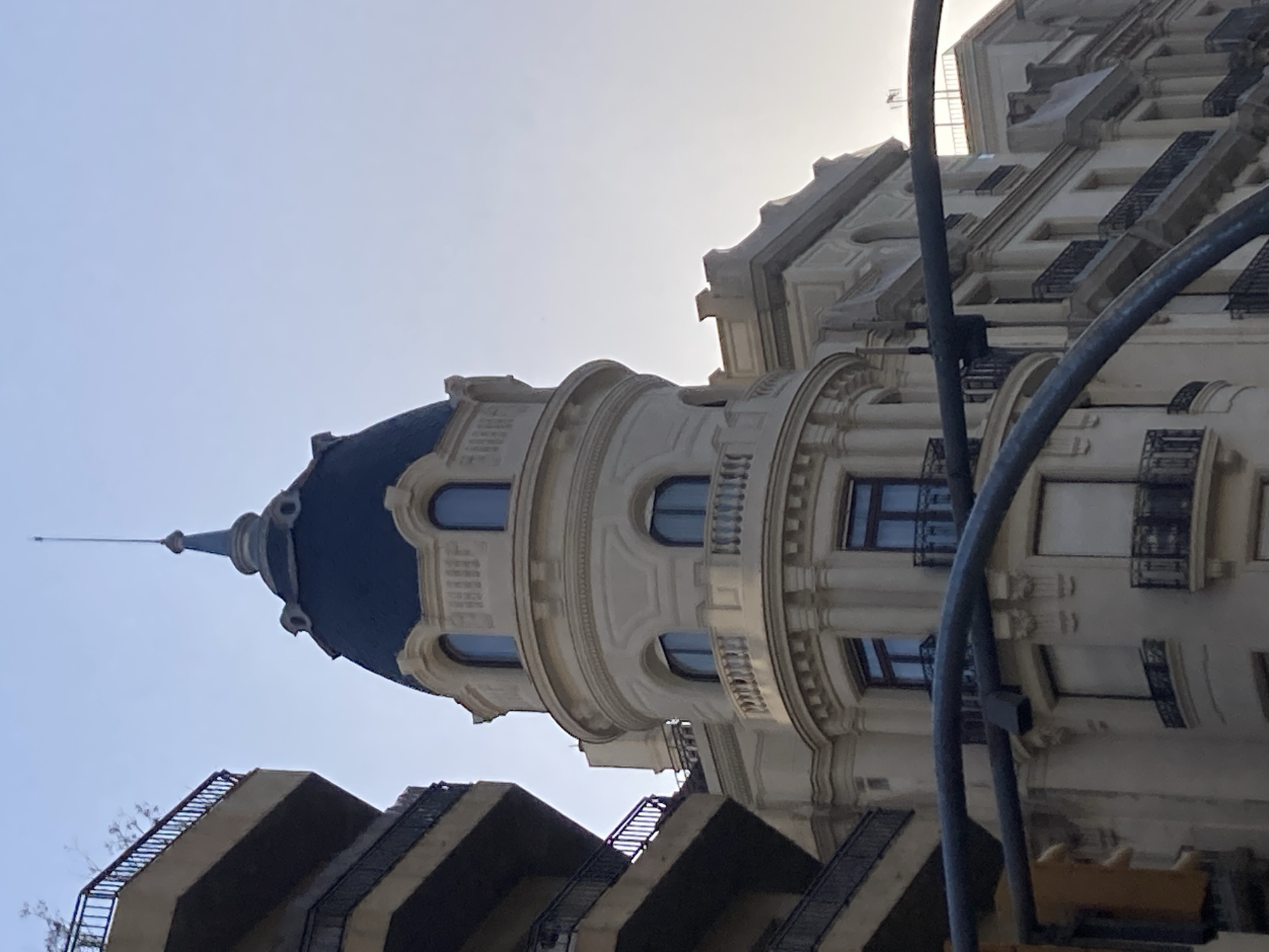 The image is an upward-facing, slightly tilted shot of a building facade with a dome and a spire. The building has a light-colored, ornate facade with multiple floors featuring balconies with black railings and arched windows. A dark blue dome caps a rounded section of the building, culminating in a thin spire that pierces the clear, pale sky. In the foreground, dark, curved pipes obscure parts of the building, suggesting they are part of a street lamp or utility pole. Below the main building, another section of architecture is visible, characterized by sharp, angular concrete structures with metal railings, possibly staircases or balconies. The scene appears to be an urban setting, likely in Barcelona, Spain, given the architectural style and the presence of similar buildings often found in the city. The light suggests it is either early morning or late afternoon, with a diffused sunlight indicating a clear sky without direct harsh light. There is no text visible in the image.
