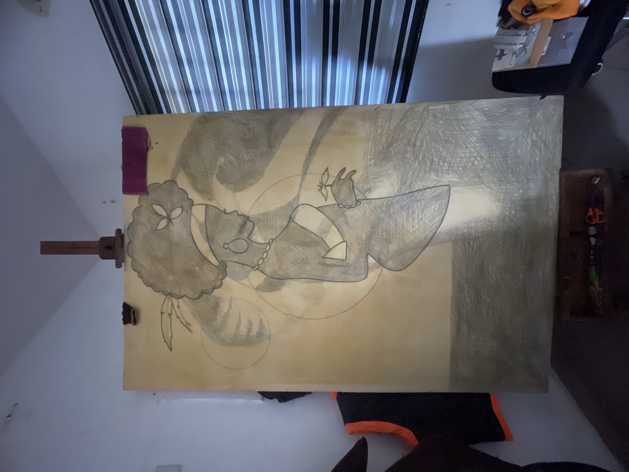 An image displays a large, vertically oriented artwork in progress, mounted on a wooden easel within an indoor setting. The artwork, appearing to be a pencil or charcoal drawing on a light brown or beige canvas, features a stylized female figure.
The figure is depicted in profile, facing left, with a substantial, textured hairstyle resembling an afro, adorned with a delicate, flower-like ornament. She wears a beaded necklace and a dress or top with a distinct collar and sleeves. Her hand is delicately holding a small, intricate, flower-like object. Faint circular and curved sketch lines are visible, suggesting the underlying composition. The drawing uses shading to create depth, particularly in the hair and clothing, while the background of the canvas is lightly textured with a similar shading technique.
In the background of the room, striped black and white vertical blinds are visible, suggesting a window. On the top left edge of the canvas, there is a small, rectangular purple object, possibly a sticky note or art supply. To the upper right, a glimpse of another object, possibly an electronic device or storage, with orange fabric is seen. In the lower right corner, a black and orange fabric item is partially visible. The overall impression is that of an artist's workspace or a room where creative work is displayed, possibly in Ado, Nigeria. No text is visible in the image. The lighting is soft and diffused.