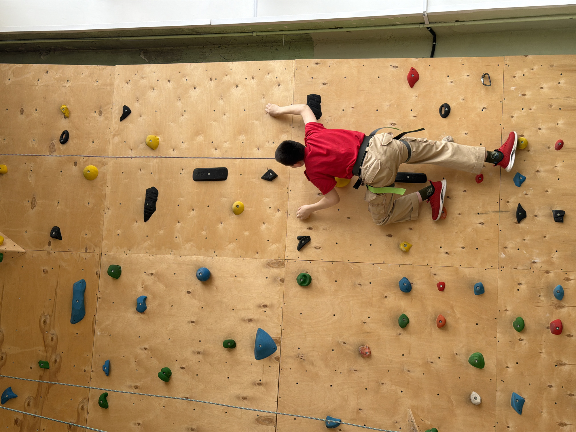 A young boy, viewed from the back, is actively engaged in indoor climbing on a wooden bouldering wall. He wears a red t-shirt, khaki cargo pants, and red sneakers, and is secured by a green climbing harness. His dark hair is cut short. The climbing wall is constructed from light-colored plywood panels, densely covered with numerous climbing holds of varying shapes and sizes, colored yellow, black, red, green, blue, orange, and white. The boy is positioned horizontally across the wall, indicating he is traversing or bouldering along a route. His hands and feet are firmly gripping the colorful holds, demonstrating focus and physical effort.
One black climbing hold on the left side of the wall distinctly displays the text "5.10," likely a brand reference. A thin, light-colored rope runs horizontally across the lower portion of the climbing wall. Above the wooden wall, a white rectangular light fixture or beam is visible, along with a greenish wall section and a black pipe or cable running along the top edge. The scene is set indoors, suggesting a climbing gym or recreational facility, with artificial lighting. The activity takes place in Izhevsk, Russia.