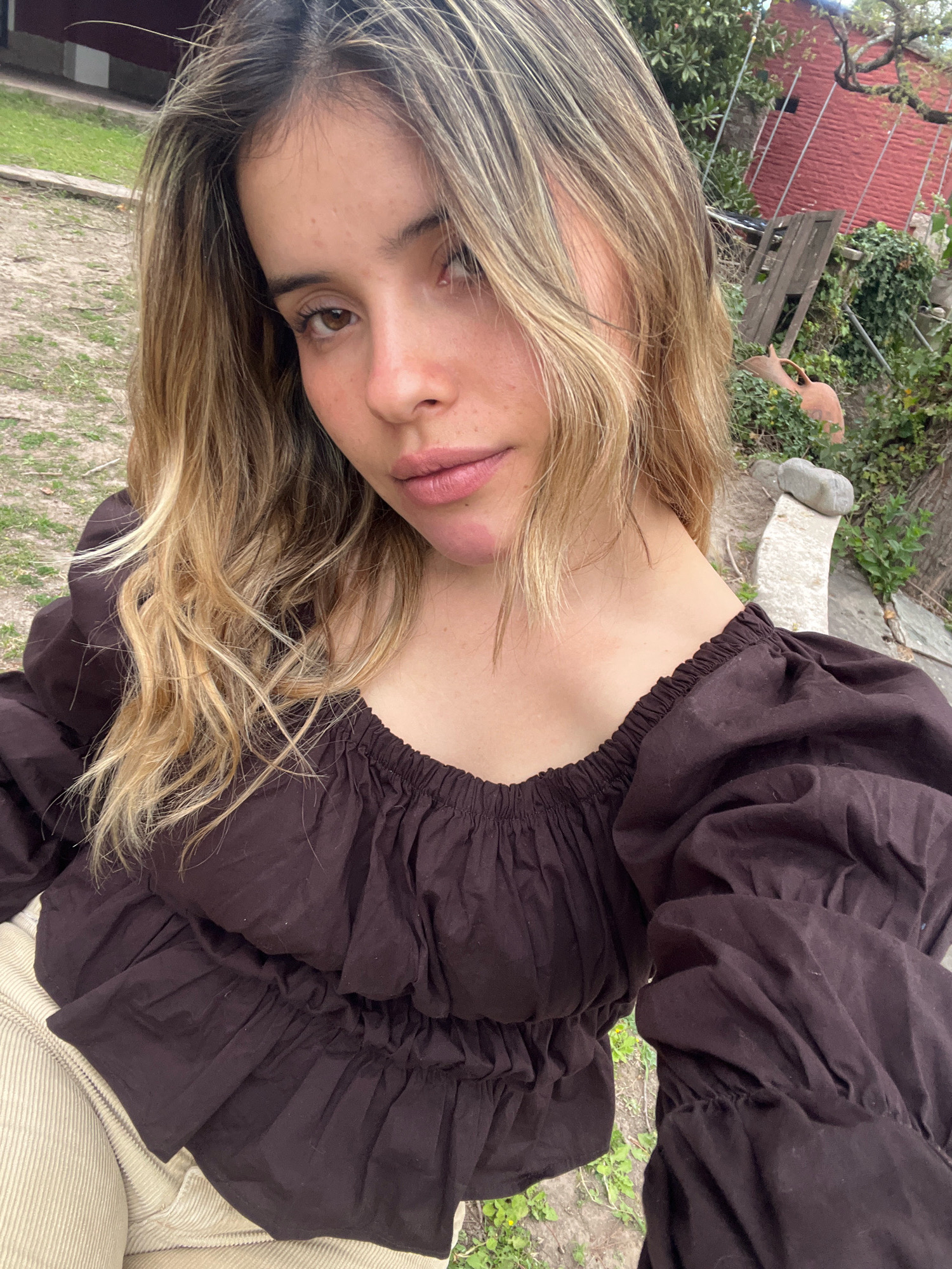 A young woman with blonde and brown ombre hair is photographed from the chest up, outdoors. She is looking directly at the camera with a neutral expression. Her skin is fair and appears to have freckles on her cheeks and nose. She is wearing a dark brown, off-the-shoulder top with voluminous puffed sleeves and ruching around the neckline and waist. Her lower half is out of focus, but she appears to be wearing light-colored corduroy pants.
The background suggests a rural or semi-rural setting, possibly a backyard or garden. To the right of the woman, there is a red brick structure, possibly a building or wall, partially obscured by greenery. Wooden planks are leaning against the wall, and an earthen jug is visible amongst the foliage. To the left of the woman, the ground is covered with dirt and sparse grass. A light-colored stone structure is also visible in the background. The overall lighting suggests it is daytime. The location is identified as El Rodeo, Argentina.