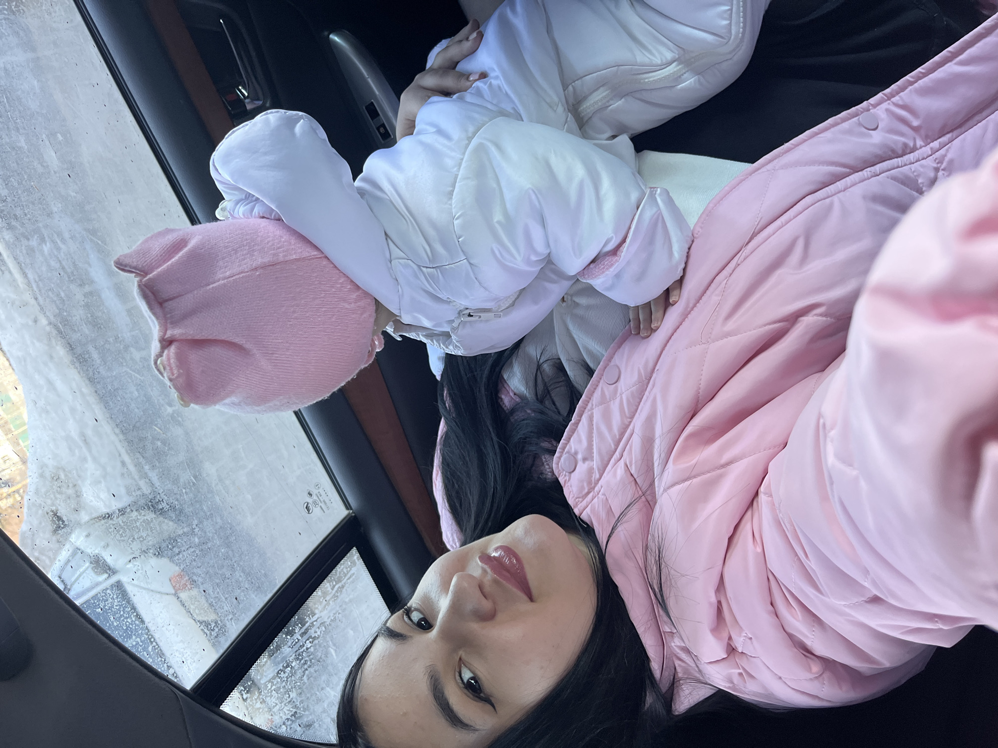 An adult female and an infant are depicted inside a vehicle in the city of Chaikovsky, Russia. The female, with long dark hair, winged eyeliner, and glossy lips, wearing a pink quilted jacket, is positioned on the left, looking directly at the camera with a slight smile. Her left hand is visible, resting on the infant's leg. The infant, wearing a white padded suit and a pink ribbed hat with ear-like features, is positioned to the right of the female, facing away from the camera. The interior of the vehicle shows a dark headliner, a sunroof above, and a dark door panel with a metallic handle and window controls. Through the wet, partially obscured sunroof, an exterior scene of snow-covered ground and faint, distant structures is visible, indicative of cold, wet weather conditions.