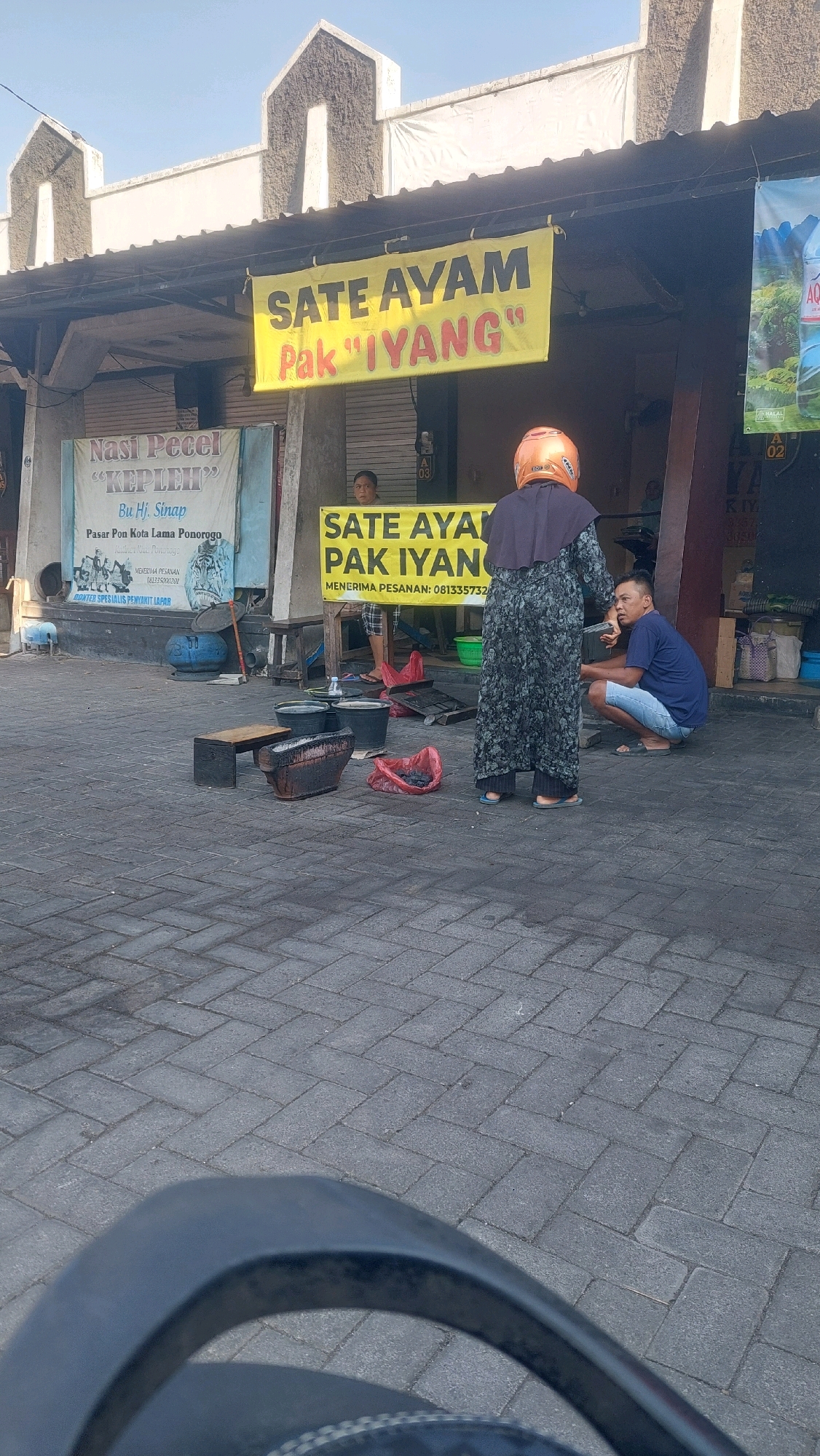 In the city of Nologaten, Indonesia, a street-side food stall is captured in an outdoor scene. A yellow banner with red and yellow text reads "SATE AYAM Pak 'IYANG'" and another similar banner is partially visible below it, suggesting the sale of chicken satay. A woman wearing a dark patterned dress and an orange helmet is standing in front of the stall, facing away from the camera. To her right, a man wearing a blue t-shirt and light blue shorts is squatting and looking towards the woman. In the foreground, there is a charcoal grill with embers, several black buckets, and red plastic bags on the brick-paved ground. A sign for "Nasi Pecel 'KEPLEH'" is visible on the left side of the stall, indicating multiple food offerings. The background shows a building with decorative elements on its facade.