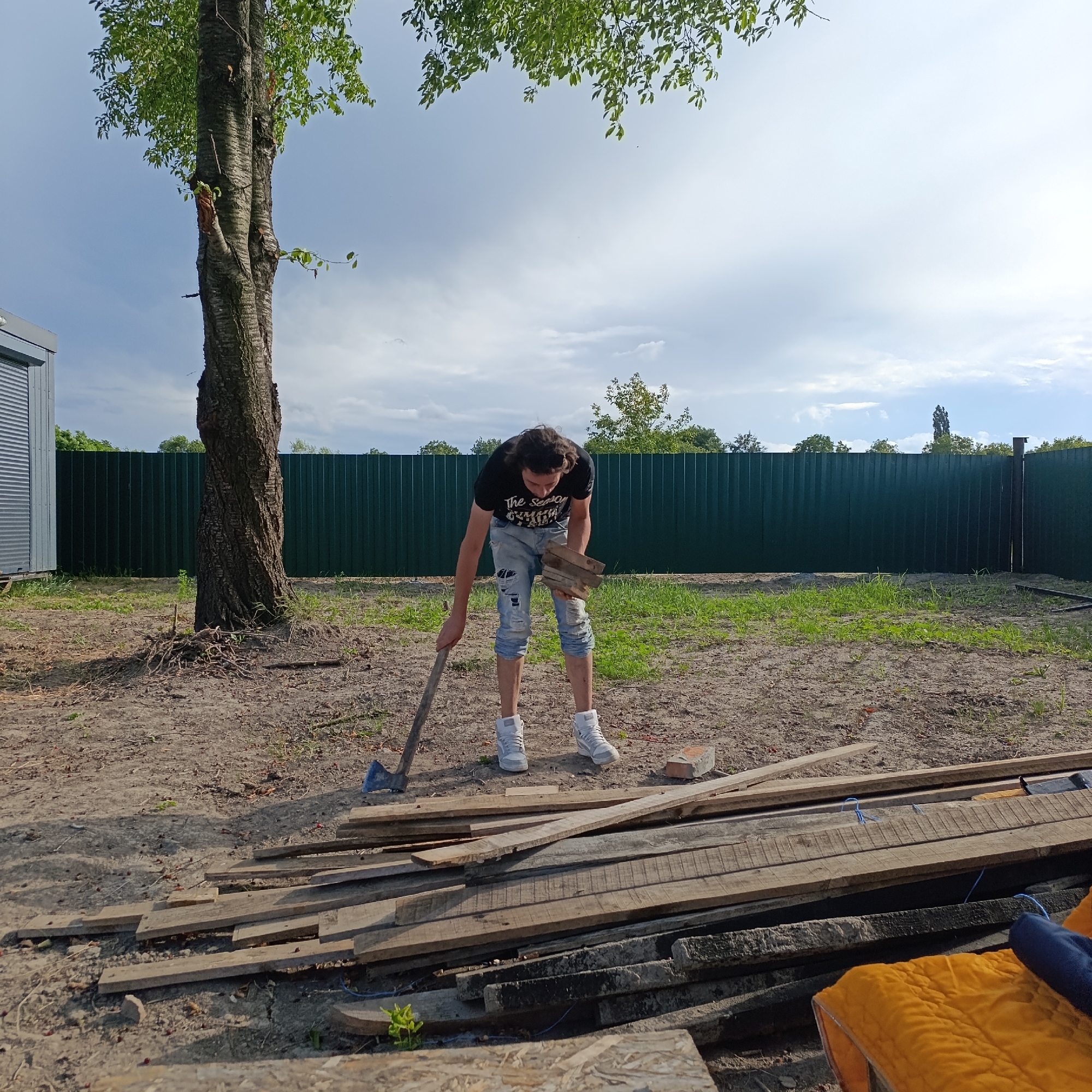 A young male with dark, curly hair is standing outside in a backyard in Ladyzhyn, Ukraine. He is wearing a black t-shirt with a graphic print and ripped blue jeans, along with white sneakers. He is bending over, holding a small stack of wooden planks in his left hand and an axe with a blue head and a wooden handle in his right hand, which is resting on the ground. In front of him, a pile of weathered wooden planks is spread across the dirt ground. Behind the man, a tall tree with a rough bark stands next to a grey building with a metal roller door. A dark green metal fence runs across the background, with trees and foliage visible beyond it under a cloudy sky. The ground is uneven and covered with dirt, scattered leaves, and some sparse green grass. In the foreground, more wooden planks are stacked, and a bright yellow object with a blue cloth draped over it is partially visible.