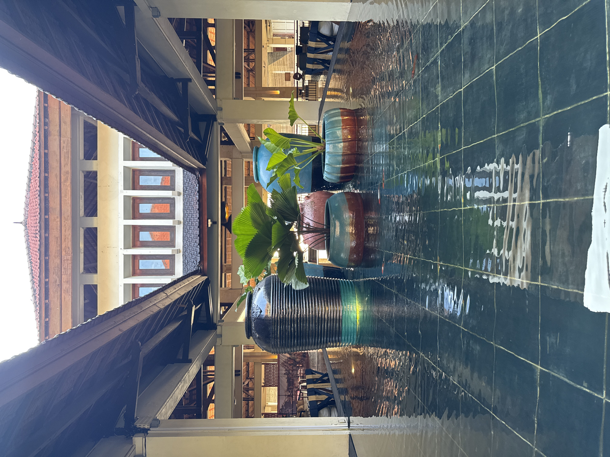 The image displays a tranquil architectural scene within what appears to be a resort or large building complex in Kuta Selatan, Indonesia. The perspective looks down a semi-enclosed corridor or walkway, featuring a prominent decorative water channel on the right side.
The water channel is lined with dark blue or green tiles, interspersed with lighter grout lines, and its surface is rippled, creating abstract reflections of the surrounding structures and light. Within this water feature are several large, decorative pots containing vibrant green plants. The pots vary in design, including a tall, dark, ridged pot; a solid blue one; and others with horizontal stripes combining blue and terracotta hues. The plants have large, broad leaves, some resembling palm fronds, adding a tropical aesthetic.
On the left side of the corridor, a substantial wooden roof structure or overhang with visible beams and rafters provides shade. Beyond this, and further in the background on both sides of the corridor, are multi-story buildings with numerous rectangular windows, some featuring wooden frames. The architecture suggests a blend of traditional and contemporary styles, common in the region.
The scene is illuminated by natural daylight, indicating it is daytime, likely morning or afternoon, given the bright reflections on the water. No specific activity or people are visible, lending a serene and peaceful atmosphere to the space. There is no visible text in the image.