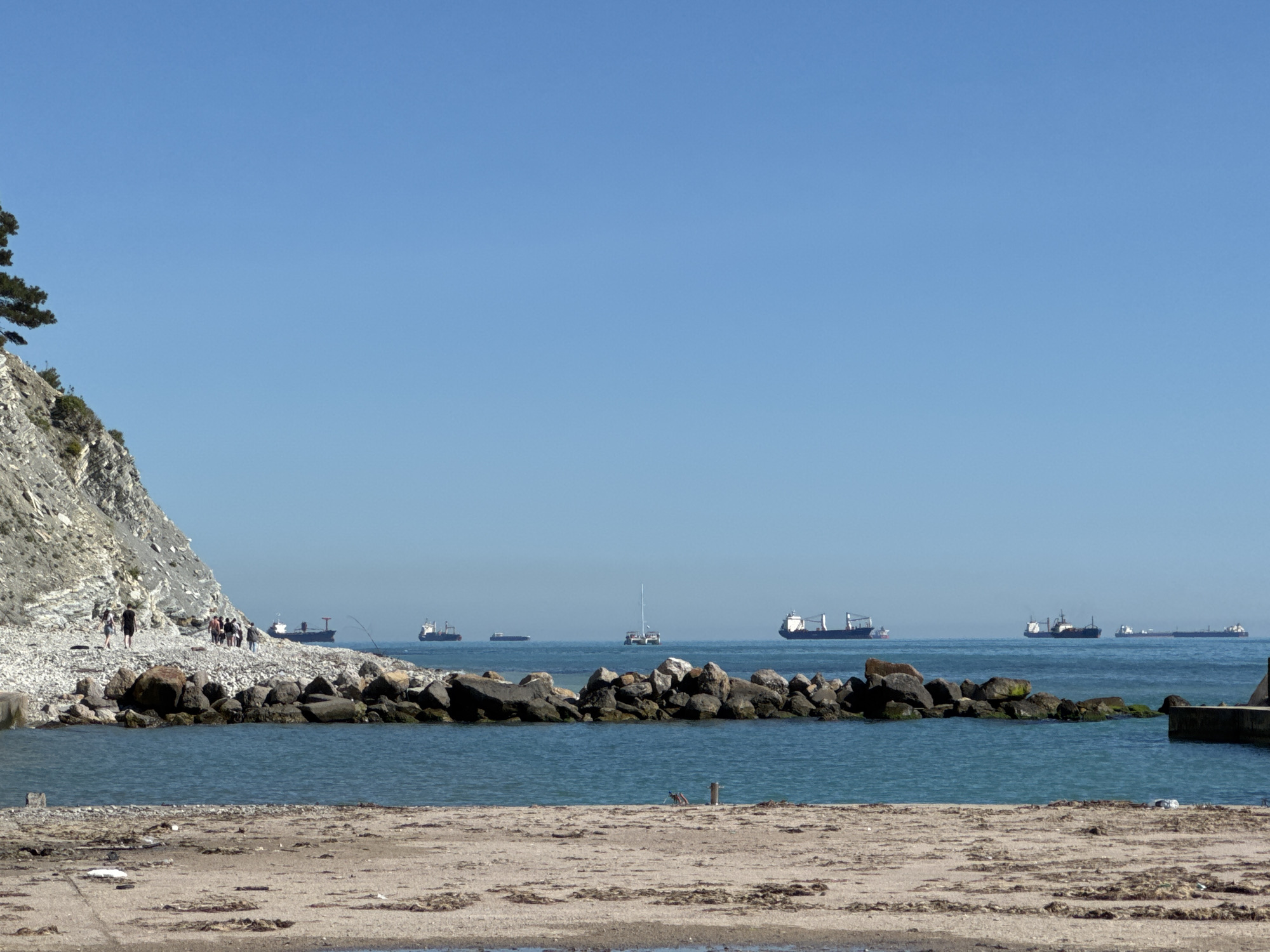 This image presents a serene coastal scene in Kabardinka, Russia, under a clear, bright blue sky. In the foreground, a sandy and pebbly beach occupies the bottom portion of the frame, showing some scattered debris or dried vegetation.
A body of calm, blue-green water fills the mid-ground, partially contained by a line of large, dark rocks forming a natural or artificial breakwater. To the left, a prominent, light-colored cliff with visible geological layers rises sharply, partially covered by sparse vegetation, with the branches of a tree extending into the top-left corner. Along the pebbly shore at the base of this cliff, a group of people can be seen walking, enjoying the coastal path. A long, slender object, possibly a fishing rod, extends into the water near the cliff edge.
In the distance, across the calm sea, multiple large cargo ships or tankers are visible, appearing to be either anchored or moving slowly. One smaller sailboat is also discernible among the larger vessels. The sky is uniformly clear and cloudless, suggesting a beautiful, sunny day. The scene combines natural beauty with the distant presence of maritime industry. There is no visible text in the image.