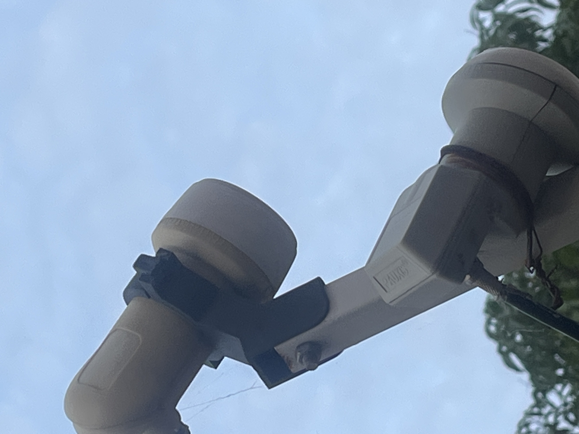 This image shows a close-up of a satellite dish LNB (Low Noise Block downconverter) against a pale blue sky. The LNB is a beige color and has a rounded end. It is attached to a metallic arm, which is also beige. A black bracket secures the LNB to the arm. There is a faint, thin thread, possibly a spiderweb, near the LNB. In the background, blurred green foliage suggests trees or bushes. A small, rectangular component with faint markings, potentially a label or model number, is visible on the metallic arm. A dark wire is attached near the bottom of the arm, with some fraying visible. The overall impression is of an outdoor, possibly residential setting, in Jalingo, Nigeria, where satellite dishes are commonly used for television reception.