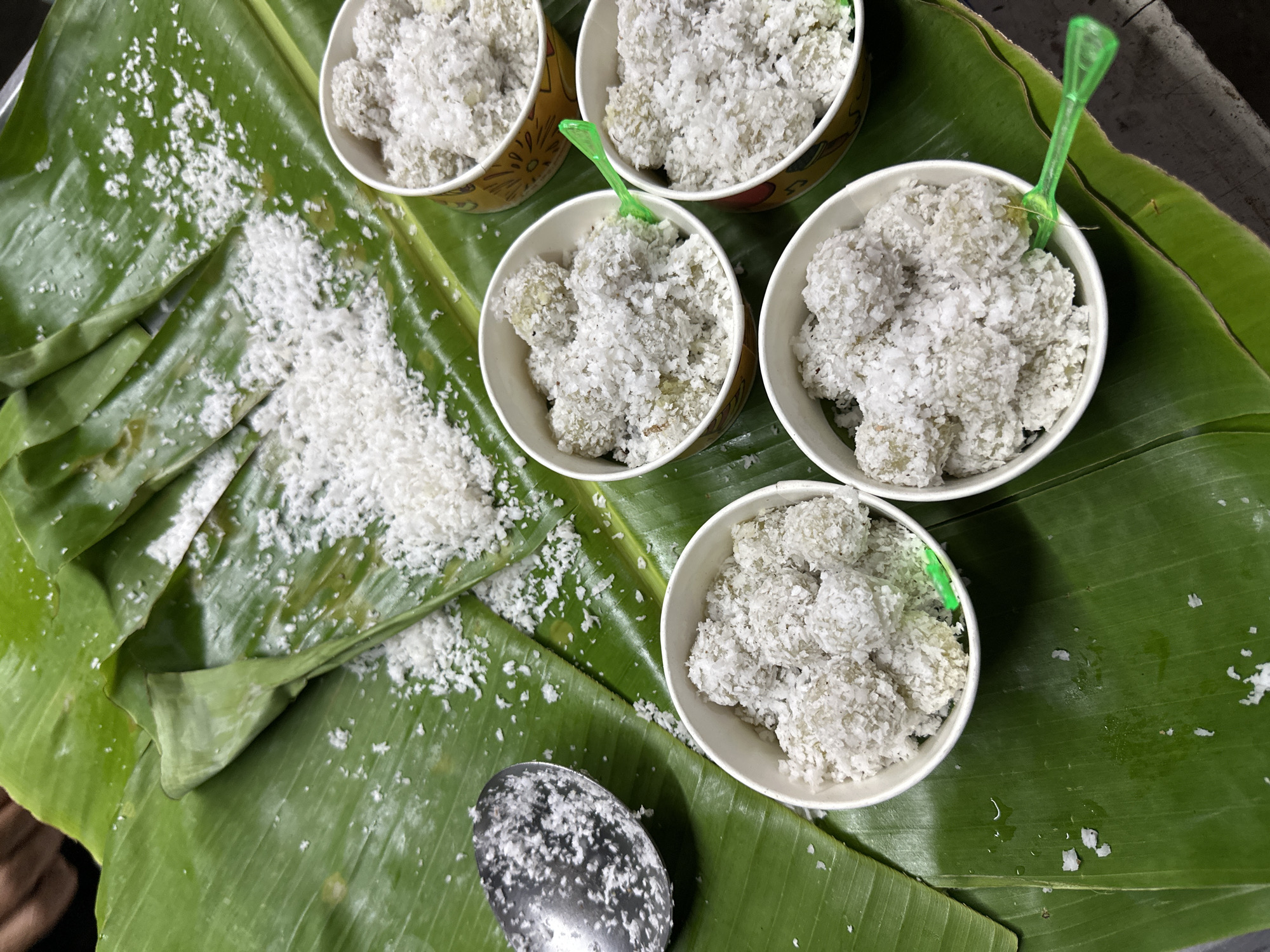 The image shows a dish called Klepon, likely in ตำบลเกาะพะงัน, Thailand. Klepon, small balls of glutinous rice flour filled with palm sugar and coated in grated coconut, are displayed in five paper cups arranged on a banana leaf. Some shredded coconut is scattered on the banana leaf. A metal spoon is also present on the leaf, covered in shredded coconut. One cup has a green plastic utensil in it. The scene seems to be a food stall or a home setting where Klepon is being prepared or served.