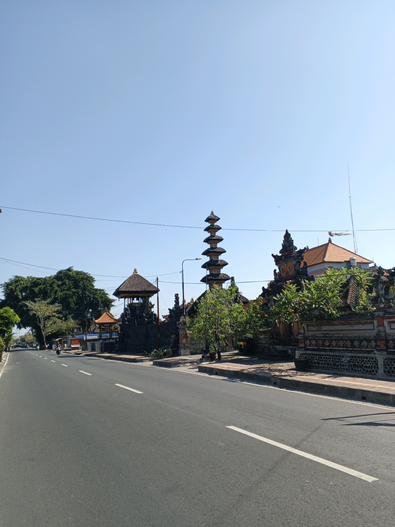 A wide asphalt road with white lane markings stretches across the foreground and into the distance on the left side of the frame. On the right side of the road, a low, ornate stone wall with intricate carvings borders a pathway and a series of traditional Balinese buildings. These structures feature tiered roofs, dark stone facades, and decorative elements. A tall, multi-tiered pagoda-like structure stands prominently among them. Lush green trees, including a large, mature tree on the left, line the roadside. The sky above is clear and bright blue. A few motorcycles and a small cart are visible on the road. The overall impression is of a street scene in a tropical, culturally rich area, likely in Abiansemal, Indonesia, given the architectural style.