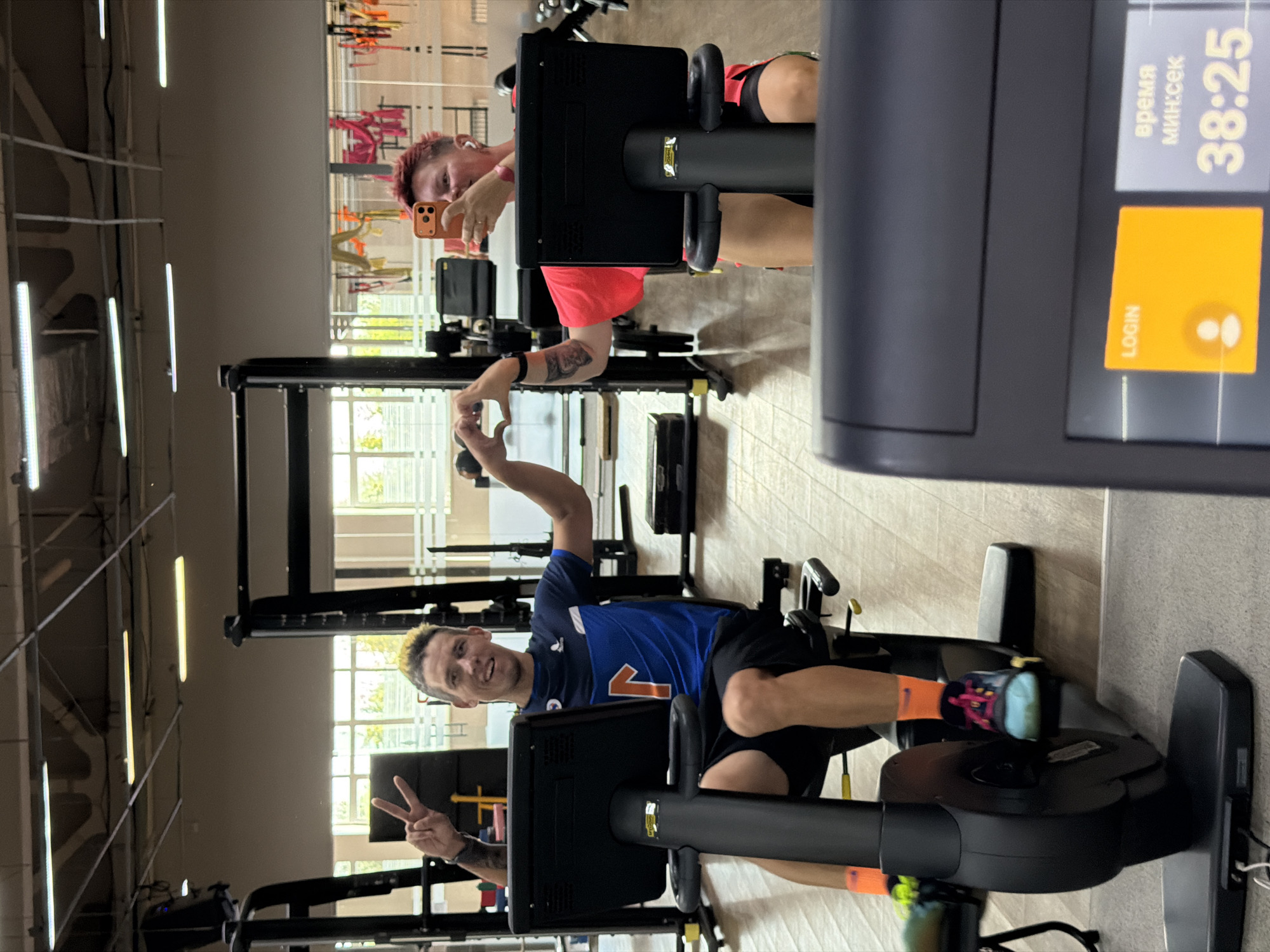 This image depicts two individuals at a gym, presumably located in Chaikovsky, Russia. The person on the left, a male with distinctive blond-streaked hair, is seated on a stationary bicycle and giving a peace sign with his left hand. He is wearing a blue and white athletic shirt and black shorts, with bright orange socks and multi-colored athletic shoes. The person on the right, with short red hair, is also on a stationary bicycle and is holding a smartphone to take a selfie, simultaneously forming a heart shape with their hands. They are wearing a bright pink athletic top and black shorts. Both individuals appear to be engaged in a workout session. The gym environment is visible, with exercise equipment, mirrors, and overhead lighting. The floor is a light-colored wood, and the walls are a neutral color. The overall atmosphere suggests a casual and active setting.