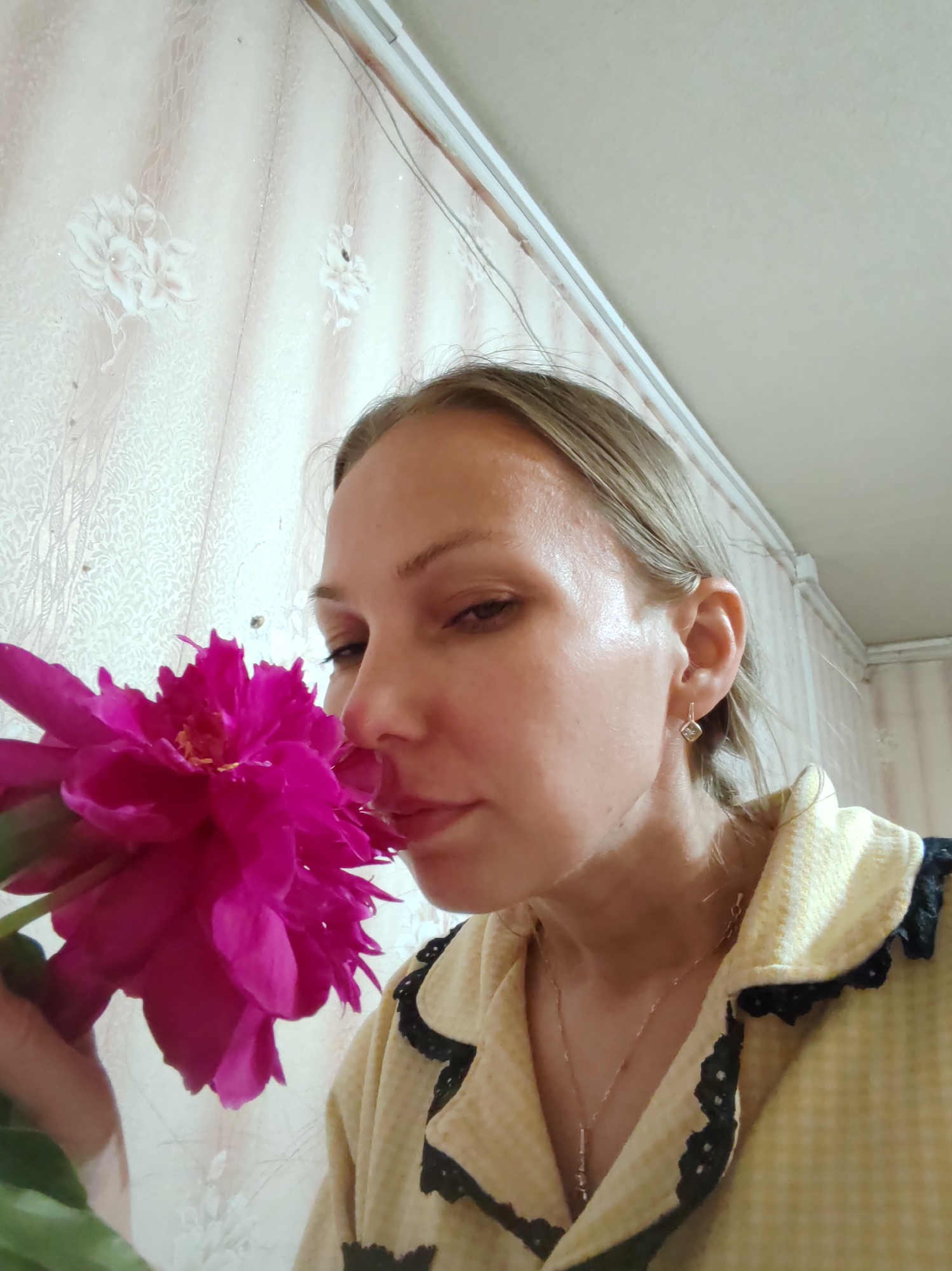 A close-up, medium shot features a woman with fair skin and light blonde hair pulled back, looking down as she smells a large, vibrant pink peony. The woman is wearing a light yellow, checkered pajama top with black lace trim on the collar and down the front. A delicate silver necklace with a small pendant rests on her chest. She is also wearing a small, diamond-shaped earring with a diamond stud in her left ear. The background consists of a pale pinkish-beige wallpaper with a subtle floral pattern. The lighting suggests it may be daytime, and the indoor setting appears to be a home in Чухлэм, Russia, as indicated by the wallpaper style. The woman's expression conveys a sense of calm and enjoyment as she savors the flower's scent. There is no visible text in the image.
