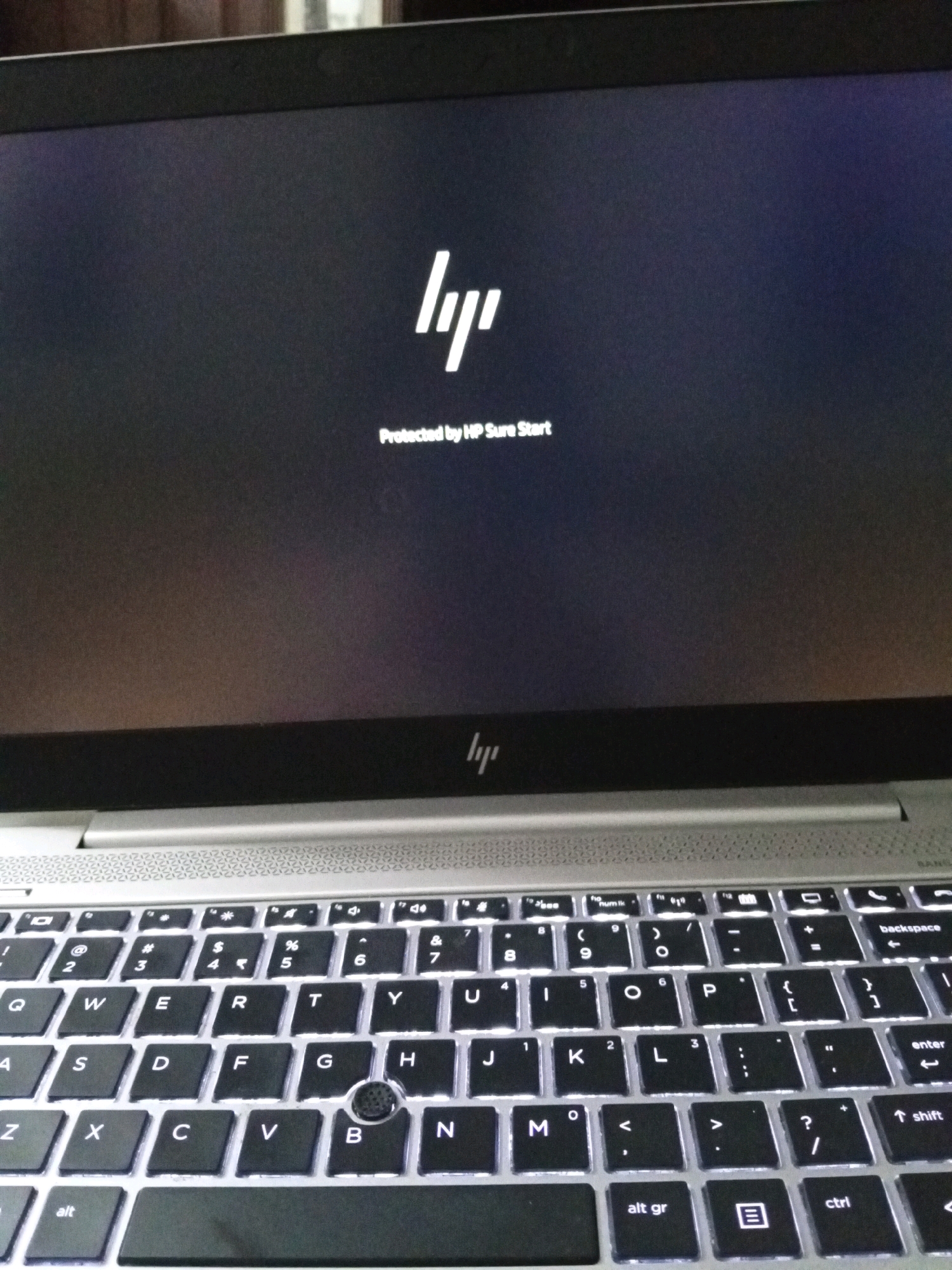 This image shows a close-up, high-angle view of an HP laptop with its screen displaying the HP logo and the text "Protected by HP Sure Start." The laptop is silver with a black keyboard that is backlit. The screen is dark, with a gradient from top to bottom. The keyboard is also visible, showing keys like 'Q', 'W', 'E', 'R', 'T', 'Y', 'U', 'A', 'S', 'D', 'F', 'G', 'H', 'J', 'K', 'L', 'Z', 'X', 'C', 'V', 'B', 'N', and 'M'. There is no indication of the city of Isma'il Pura, Pakistan, or any people or actions within the image.