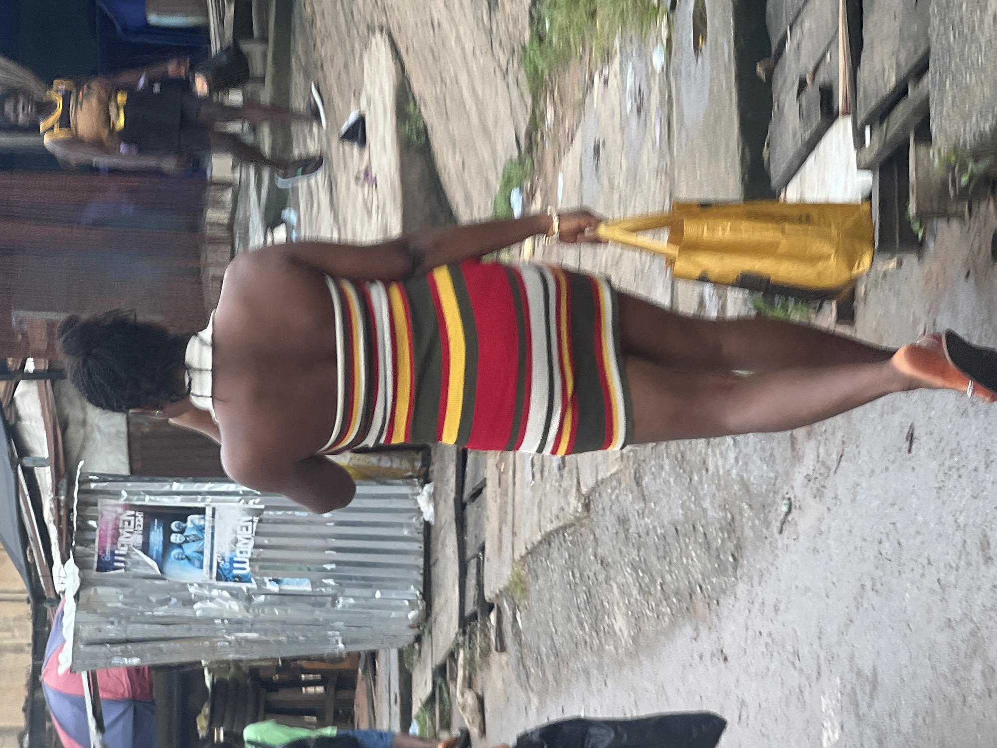 In Nkpor, Nigeria, a woman with dark skin and braided hair walks away from the camera in a striped, strapless dress and orange heeled sandals. She carries a yellow bag in her right hand and wears a gold bracelet on her left wrist. The scene takes place on a wet, unpaved street lined with corrugated metal structures and wooden planks. A poster featuring the word "WOMEN" is visible on one of the metal structures. Another woman, wearing a yellow and black outfit and dark shorts, walks in the background. The overall environment suggests a bustling urban setting with elements of informal commerce.