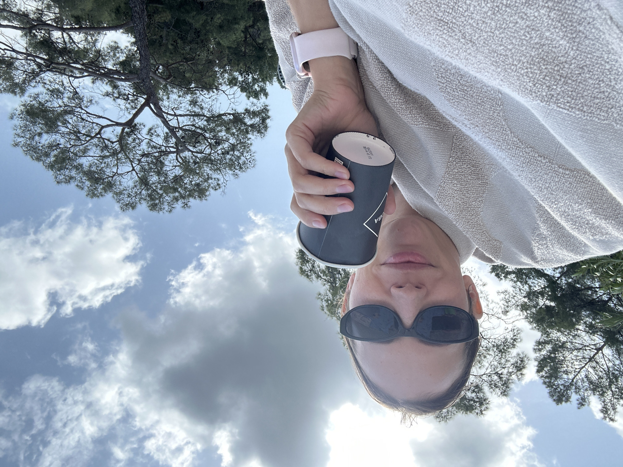 This image captures a person's upward-gazing perspective, likely a selfie, set outdoors in Fethiye, Turkey. The central subject, whose face is partially obscured by dark sunglasses, has visible lips indicating a neutral expression. They wear a light-colored, textured garment, possibly a terry cloth top, and a light pink smartwatch is visible on their left wrist. In their right hand, they hold a dark-colored, disposable coffee cup, on which the text "5A" and some smaller, indistinct writing are legible.
The background features a vibrant blue sky, partially covered by scattered white and grey clouds, suggesting daylight hours with mixed weather. Overhead, large trees with dense green foliage and intricate branch patterns frame the sky, characteristic of Mediterranean pines found in the region. The overall scene conveys a serene moment spent enjoying a beverage in a natural outdoor environment.