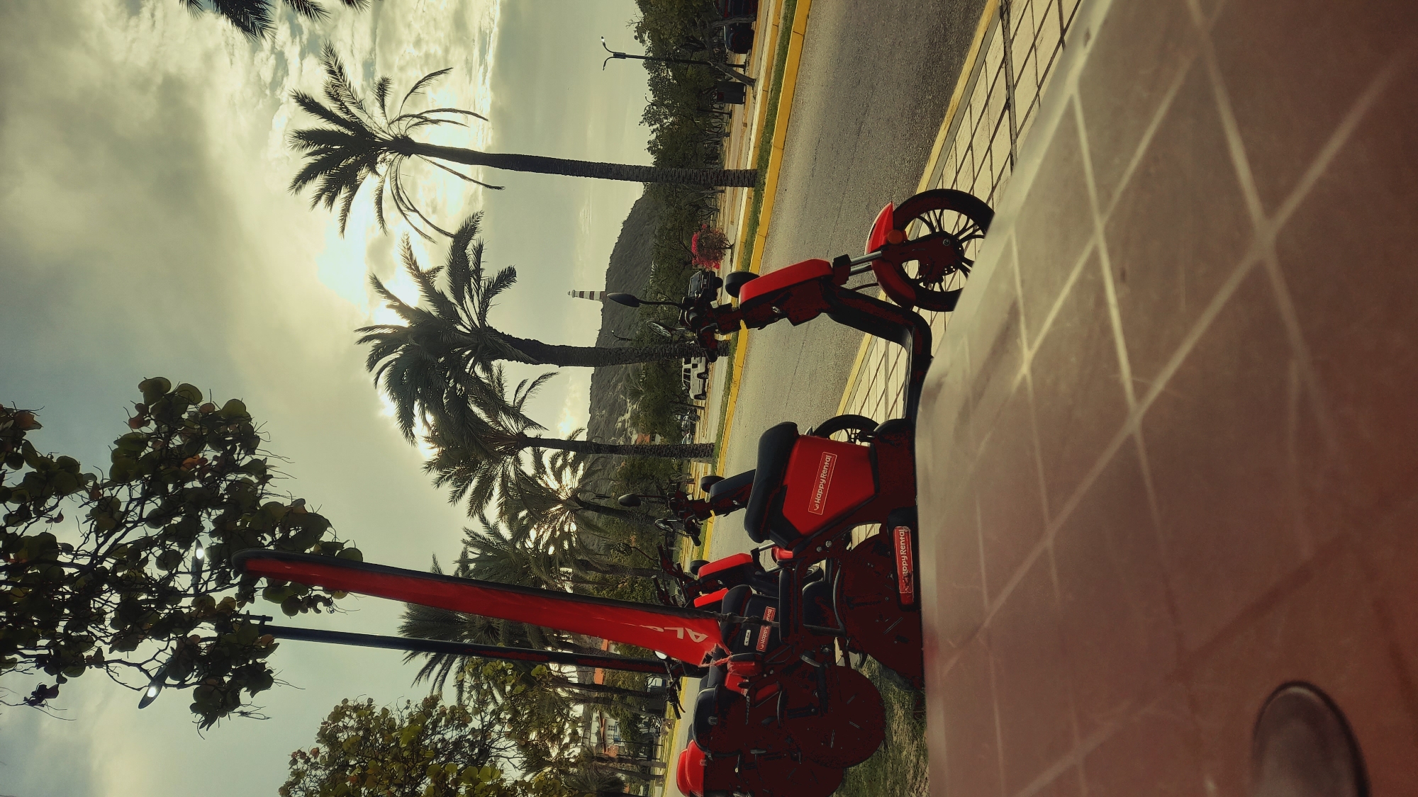In the city of Pedro González, Venezuela, a scene unfolds with several red electric scooters lined up along a sidewalk next to a road. The scooters are modern and appear to be part of a rental service, as indicated by the "Happy Rental" logo visible on some of them. In the background, palm trees dominate the landscape, with lush green foliage and their characteristic slender trunks reaching towards a cloudy sky. The sun is breaking through the clouds, casting a soft, diffused light on the scene. Beyond the palm trees, a gentle slope rises, topped by a lighthouse, hinting at a coastal or near-coastal location. The road is paved and has yellow markings, with a few vehicles visible in the distance. The foreground is occupied by a textured surface, possibly a table or a tiled area, which is out of focus, drawing attention to the scooters and the environment. The overall impression is one of a tropical setting, likely a tourist area, where these electric scooters are available for rent.