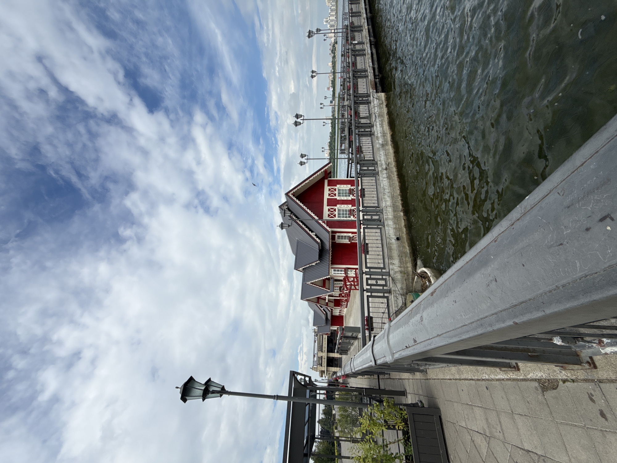 The image shows a scenic view of a waterfront promenade in Kazan, Russia. In the foreground, a grey metal railing lines a paved walkway. Beyond the walkway, a body of dark, rippling water stretches out. On the water, a pier extends outwards, lined with decorative streetlights and a series of charming, red buildings with white trim and dark roofs. These buildings appear to be part of a restaurant or cafe complex, designed in a traditional architectural style. The sky above is partly cloudy, with patches of blue sky peeking through fluffy white clouds. In the distance, the city skyline of Kazan is visible, with modern buildings contrasting with the historic waterfront structures. The scene is peaceful and evokes a sense of leisure and enjoyment of the urban waterfront.