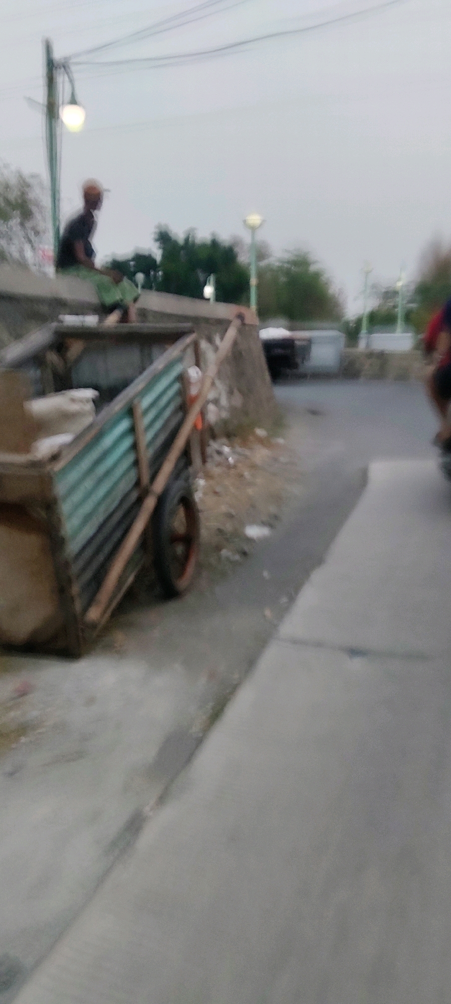 A medium shot captures an outdoor scene in Pasirsari, Indonesia, with a low-angle perspective. The foreground is dominated by a weathered wooden cart with a corrugated metal siding. The cart is loaded with unseen contents, partially covered by light-colored material. A single wheel, with a reddish-brown rim and black tire, is visible on the right side of the cart. The cart is situated on an asphalt surface that transitions to a paved walkway.
In the mid-ground, a person wearing a dark shirt and a light-colored cap is seated on a concrete ledge, facing left and looking downwards. Behind the seated person, a street light with a glowing bulb is mounted on a green pole. Further back, trees and more street lights are visible, suggesting a public area. The background is a hazy sky with overhead power lines. To the right of the cart, a motorcycle with a rider is partially visible on the road, moving away from the camera. The overall ambiance is muted due to the overcast sky.