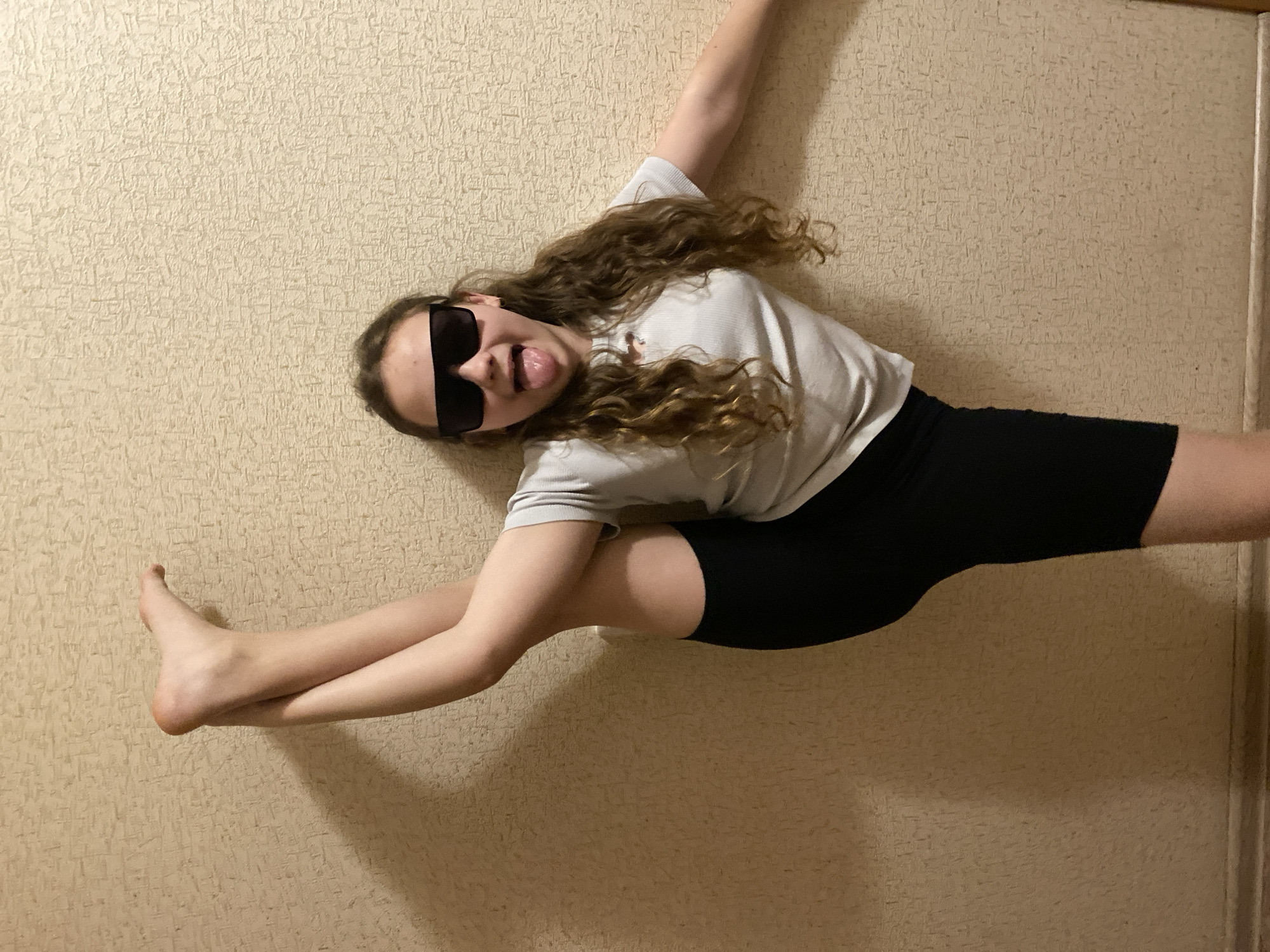 A young woman is performing a split against a textured, beige wall. She is wearing black athletic shorts and a white t-shirt. Her dark sunglasses obscure her eyes, and she is sticking her tongue out. Her curly hair is light brown. One arm is extended upwards towards the top left corner of the frame, and the other is bent near her torso. The location is Unknown, Unknown.