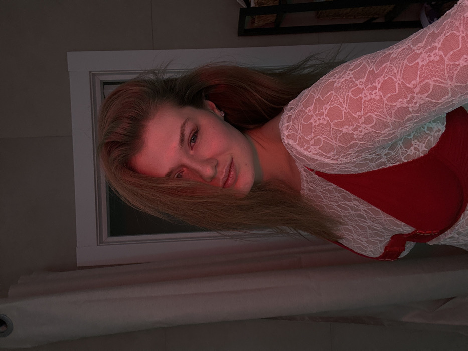 A young woman with long blonde hair, illuminated by a red light, is lying down. She is wearing a red bra covered by a white lace top. The background is blurred, showing a white frame around what appears to be a window, and the bottom is draped with white fabric. Her hair partially covers her face, and she has a slight smile. A black shelf with objects is visible in the background.