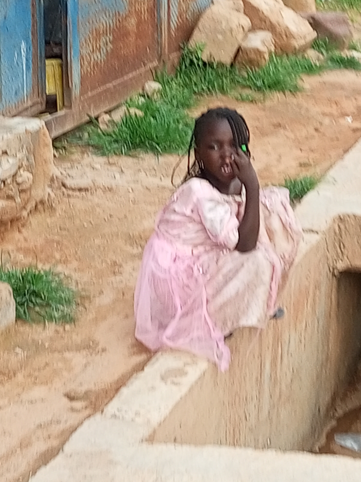 A young girl with braided dark hair and a green bead in her hair sits on a concrete ledge. She wears a light pink dress. Her right hand is raised to her mouth, and she appears to be looking at the camera. The girl is in an outdoor setting with dirt and rocks visible. Green grass grows in patches around her. Behind her is a weathered, rusty metal structure with a blue panel. The overall impression is of a humble, outdoor environment. The time of day appears to be daytime, with natural light illuminating the scene. There are no visible signs of text in the image. The setting suggests a rural or underdeveloped area, consistent with the provided location context of Tempure, Nigeria.