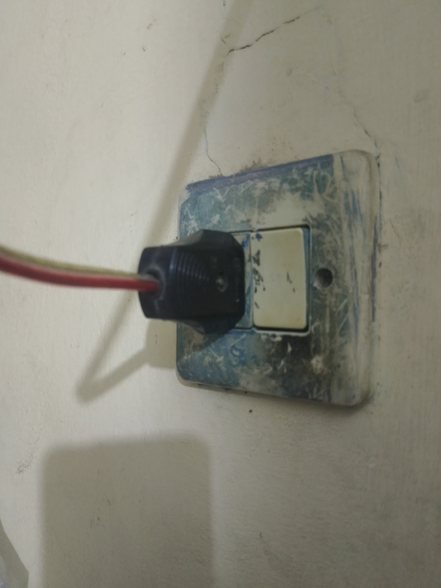 A black electrical plug with red and yellow wires extending from it is inserted into a light-colored electrical outlet mounted on a textured, off-white wall. The outlet cover appears to be a bluish-grey and shows signs of wear and possibly some scuff marks or dirt. A light switch is visible on the outlet cover. The wall has a few visible cracks and imperfections. The lighting suggests an indoor environment. The image was captured on a wall in Mukapayung, Indonesia.