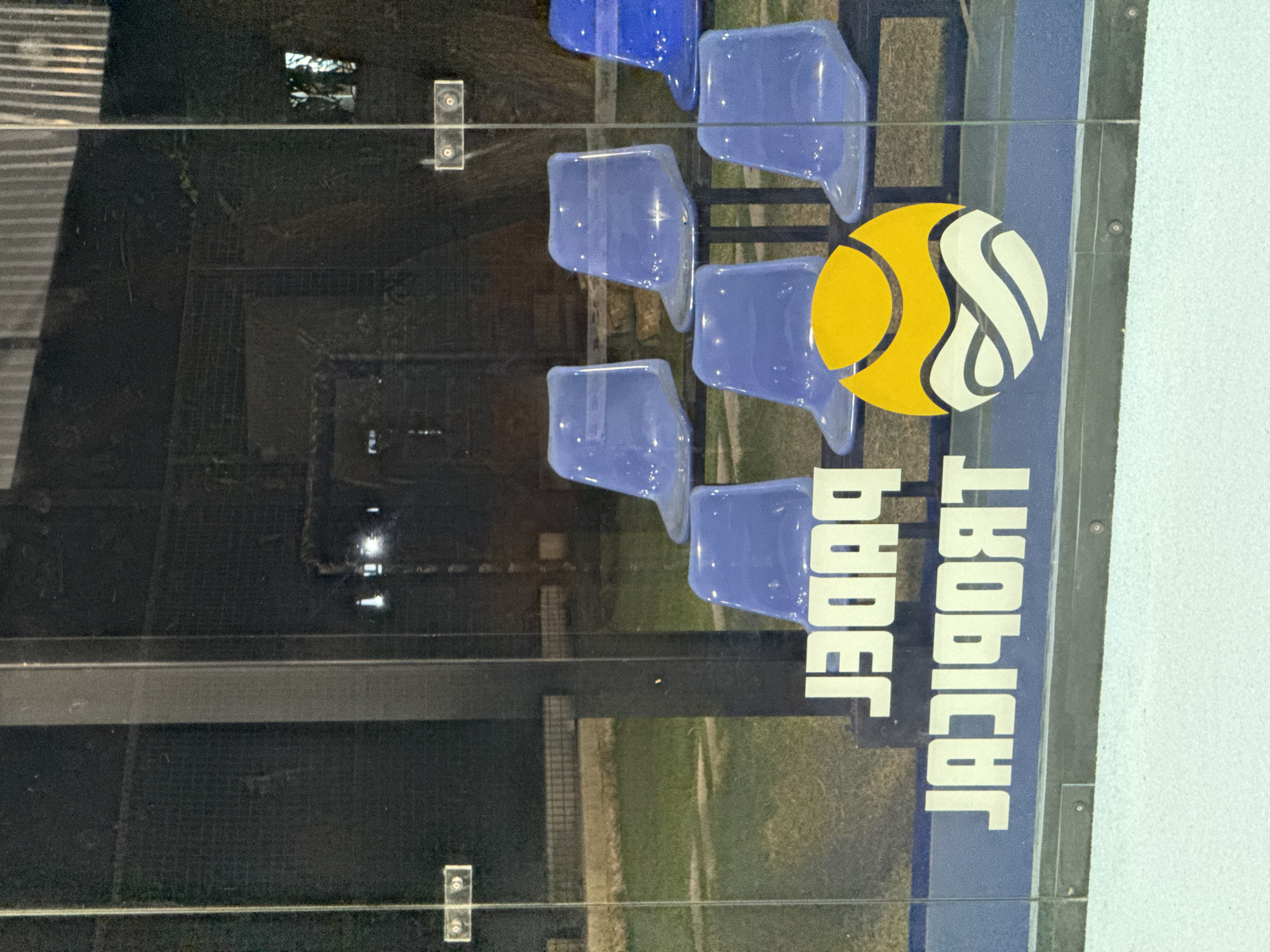 This media file depicts an outdoor setting, likely a sports venue, with rows of empty blue plastic stadium seats. The seats are arranged in a staggered pattern, suggesting a seating chart for spectators. Reflections are visible on the glossy surface of the seats and a dark barrier, obscuring details of the surrounding environment.
A prominent graphic featuring a yellow circle with white stylized curves is visible, resembling a tennis ball or a similar sports emblem. Below this graphic, white text is displayed vertically on a blue background. The text appears to be reversed, reading "JABOIBOT" and "BBOER" if viewed correctly. The overall impression is of a sports facility at dusk or night, given the artificial lighting suggested by reflections. The weather appears clear, and the location context suggests it is in Baan Bang Rak, Thailand. No people or specific activities are depicted.
