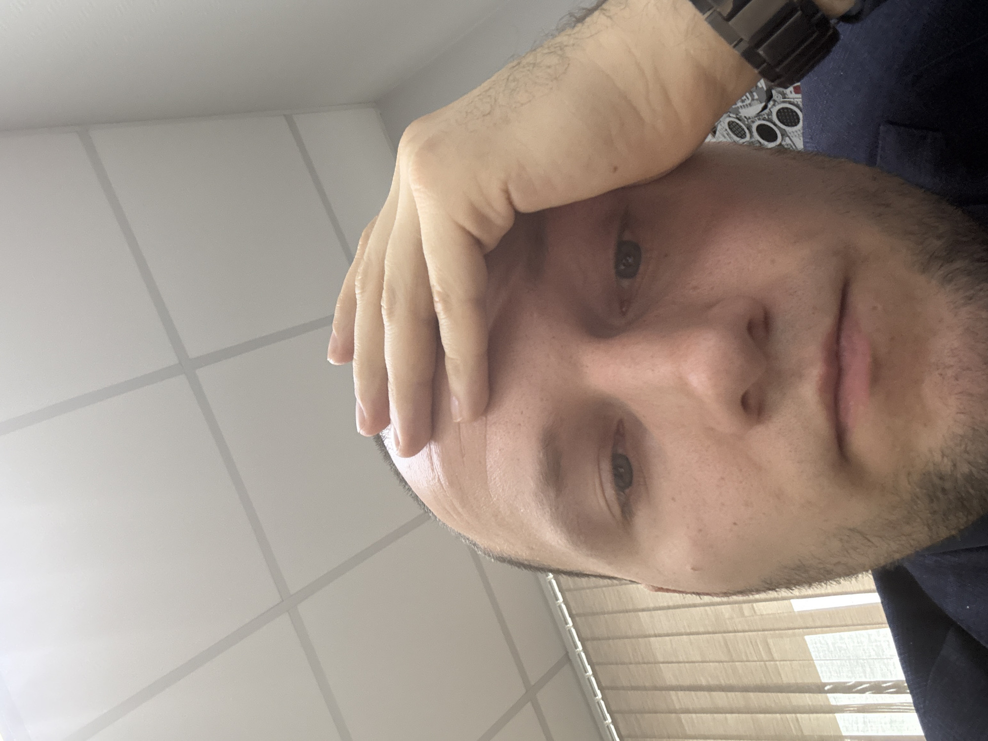 A close-up, self-shot image of a man indoors. He has fair skin, dark hair, and a short beard. His right hand is raised and partially obscures his forehead. He is wearing a dark-colored jacket and a watch. The background consists of a white ceiling with grid lines and a window with closed blinds. There's no information to suggest that the location is Krasnoyarsk, Russia.