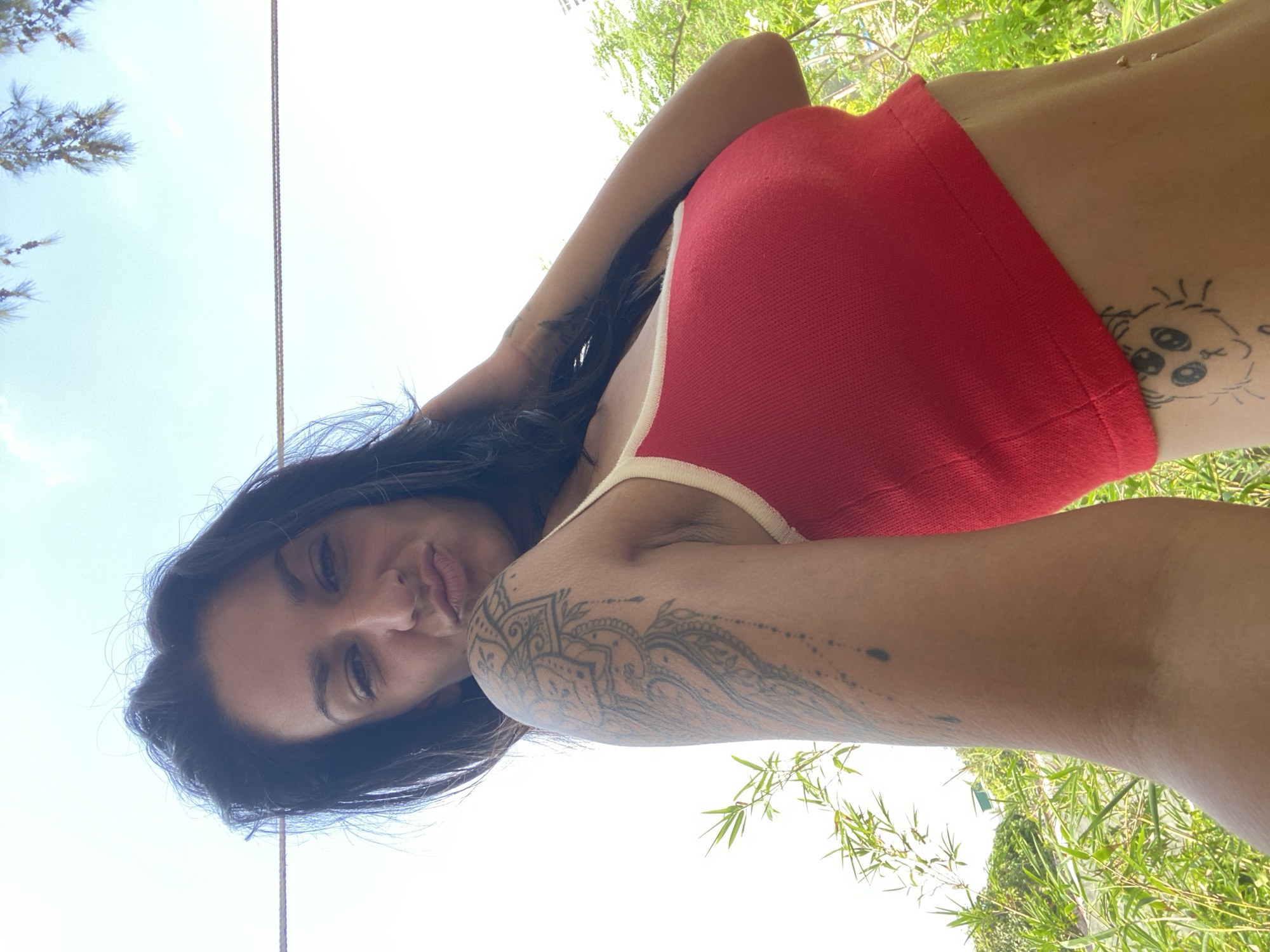 A young woman with dark hair and tattoos on her arms and torso is posing for a selfie outdoors. She is wearing a red crop top with a white trim. The background shows greenery and a bright sky. The image appears to have been taken in Mũi Né, Vietnam, given the tropical vegetation and bright sunlight characteristic of the area.