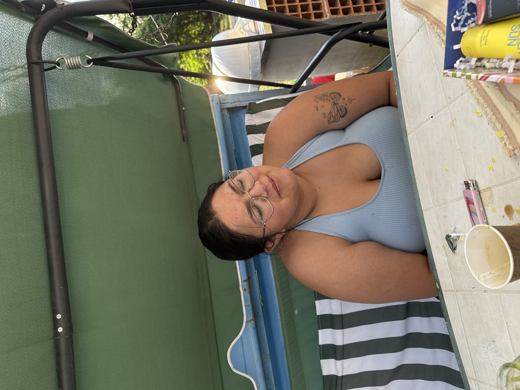 A woman with dark hair and glasses is lying on her side, looking up towards the camera. She is wearing a light blue ribbed tank top. A butterfly tattoo is visible on her upper arm. She appears to be relaxing outdoors on a swing or lounge chair, with a green canopy overhead. In the background, there is a striped cushion, some greenery, and a white structure. A table is visible to the right, with a paper cup, a pink lighter, and a yellow bottle of sunscreen on it. Some yellow crumbs are scattered on the table. The scene suggests a warm, sunny day, likely in the afternoon, given the lighting. The overall mood is relaxed and casual. The yellow bottle of sunscreen has visible text that includes "SUN". The location is identified as Sredets, Bulgaria.