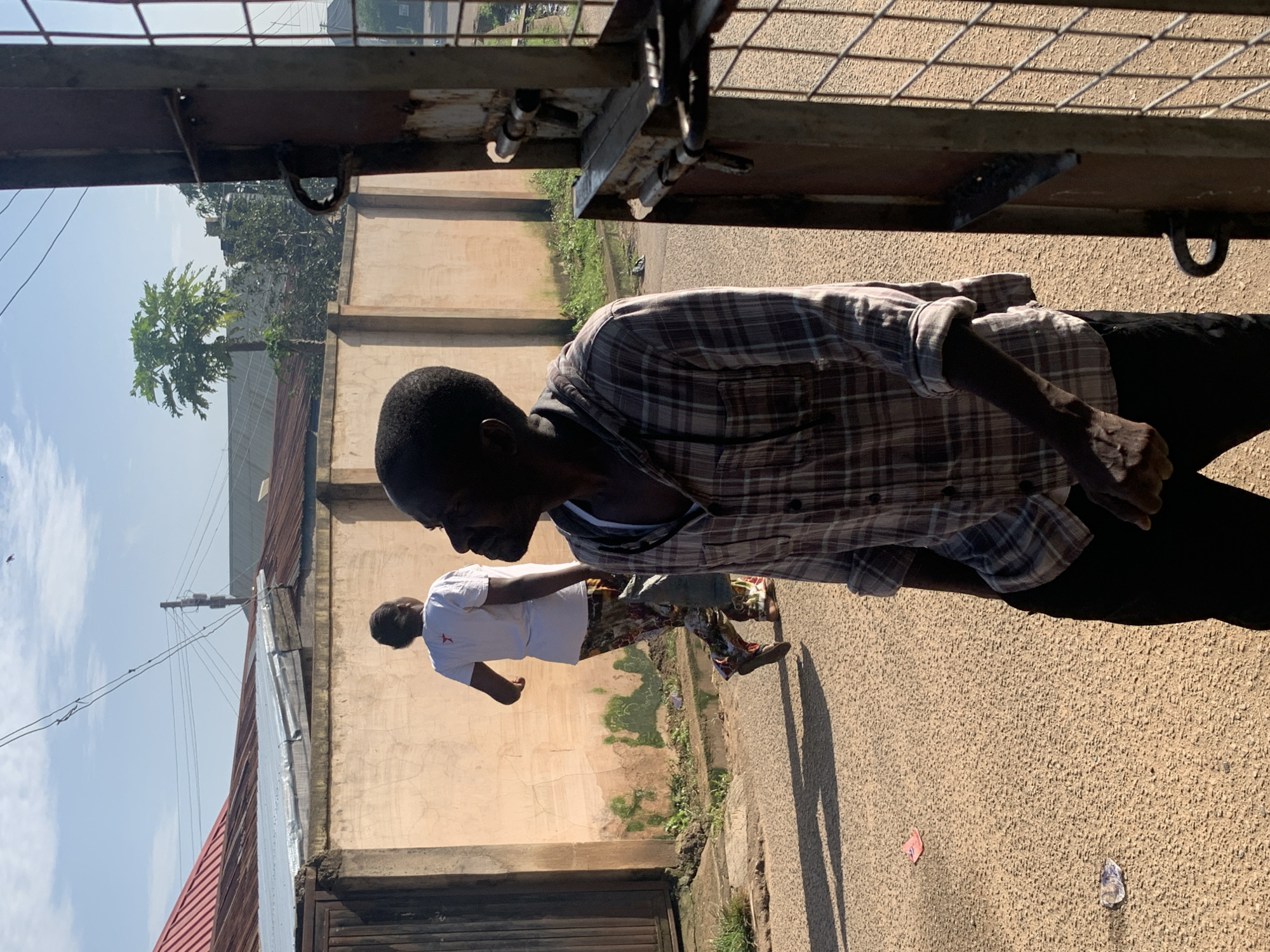 A man wearing a plaid shirt and dark pants stands in the foreground, looking to the right. Behind him, a person wearing a white t-shirt with a red star emblem and patterned pants walks away from the camera. They are on a paved road in what appears to be a residential area. A textured, pale wall runs along the side of the road, with some green moss or growth near the bottom. Buildings with corrugated metal roofs are visible in the background. A rusty metal gate with a latch is in the right foreground. The scene is illuminated by bright sunlight, casting shadows on the ground. The image is taken in Jos, Nigeria.