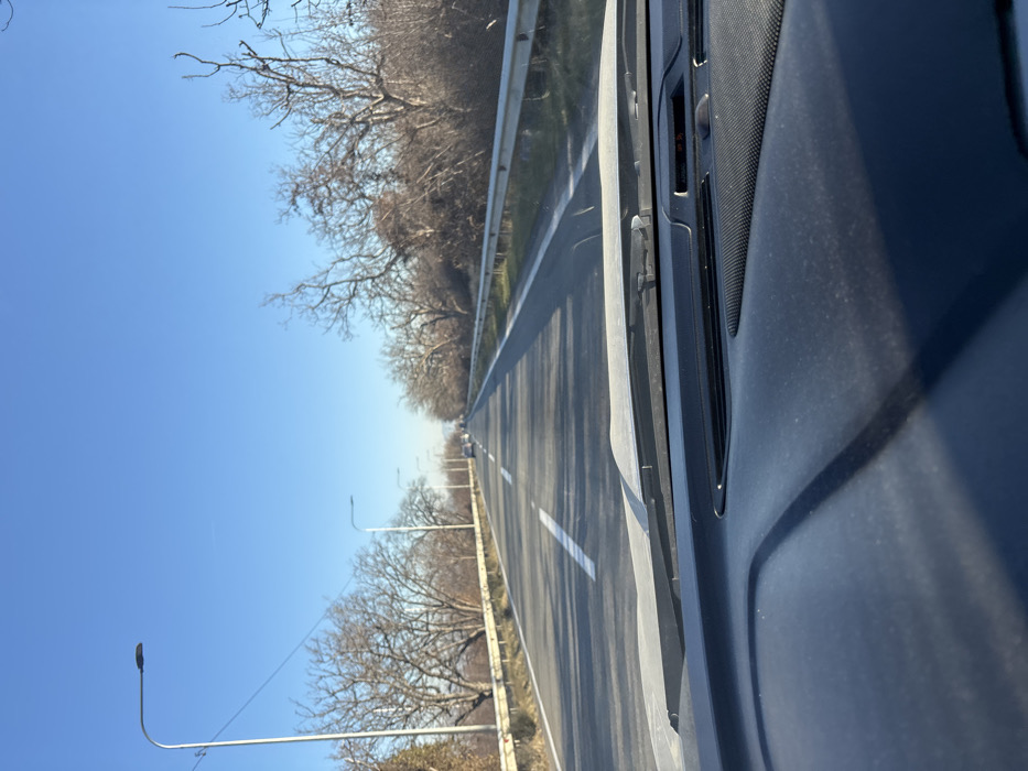 The image displays a two-lane paved road in Changilari, Georgia, viewed from the interior of a vehicle. The foreground exhibits a dark, dust-covered dashboard and the lower portion of the windshield, including a wiper blade. The road features white lane markings, with a broken centerline and a solid white line defining the right edge. Metal guardrails run along both sides of the road. Beyond these guardrails, numerous bare deciduous trees and dense brush line the path, suggesting a dormant season. Multiple streetlights with dark, elongated fixtures are spaced along the left side of the road, receding into the distance. Several vehicles are visible traveling in the same direction in the far distance. A clear, bright blue sky occupies the upper frame, with a faint, hazy outline of distant landforms visible on the horizon. The scene is illuminated by direct sunlight.