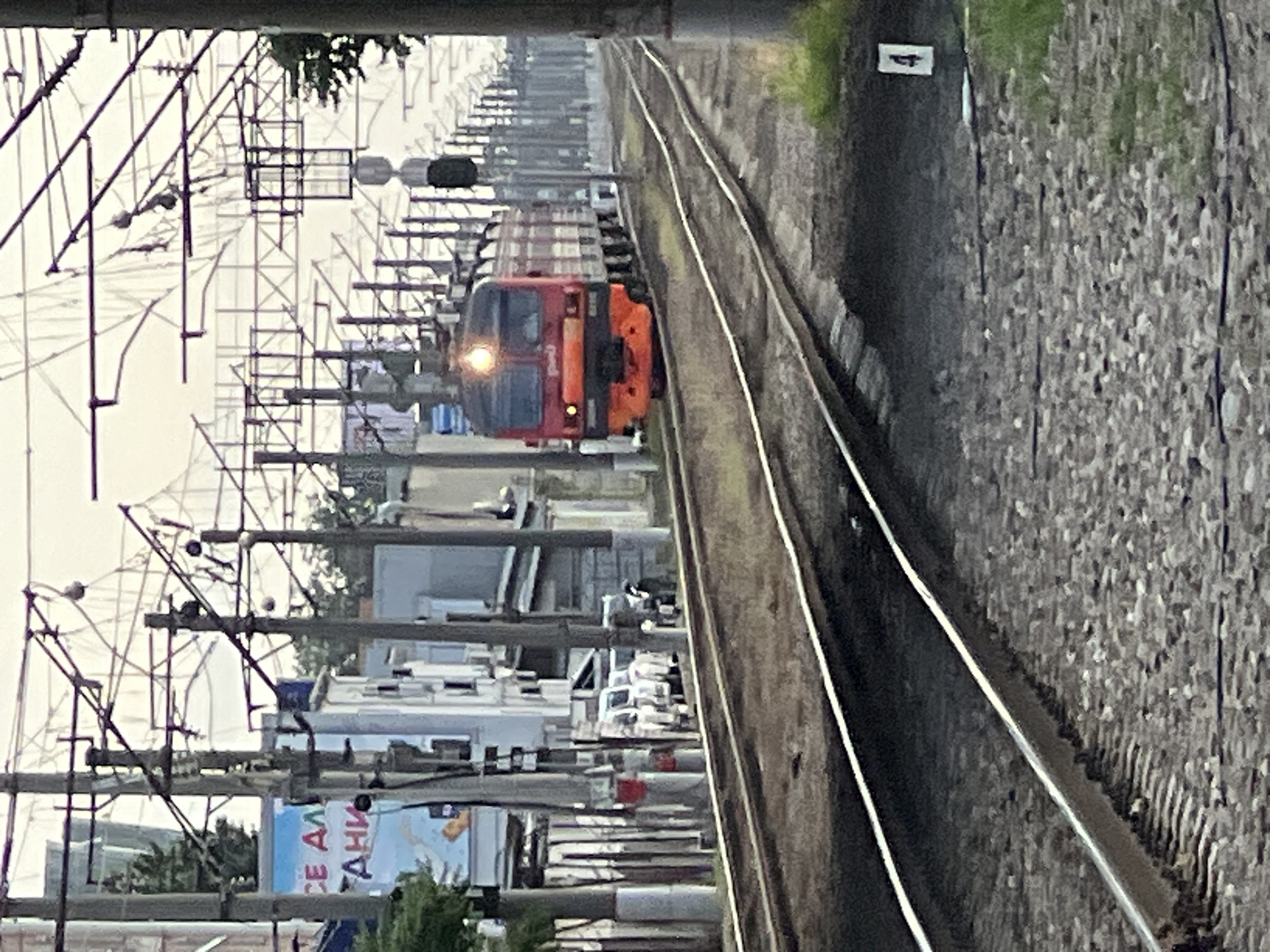 A red and orange electric train is approaching on a set of railroad tracks. The train has several cars, and its headlight is illuminated. The tracks are flanked by a gravel bed and some sparse green vegetation. Overhead, there are numerous electrical wires and poles, characteristic of a train station or railway line. In the background, there are buildings, some of which appear to be commercial or industrial, with signage visible. A few parked vehicles, including what appear to be white vans or minivans, are visible along the side of the road or track. A traffic light or signal is also present near the train. The sky is overcast. The scene is consistent with the railway infrastructure found in Krasnodar, Russia, likely near a station or along a major transit route.
