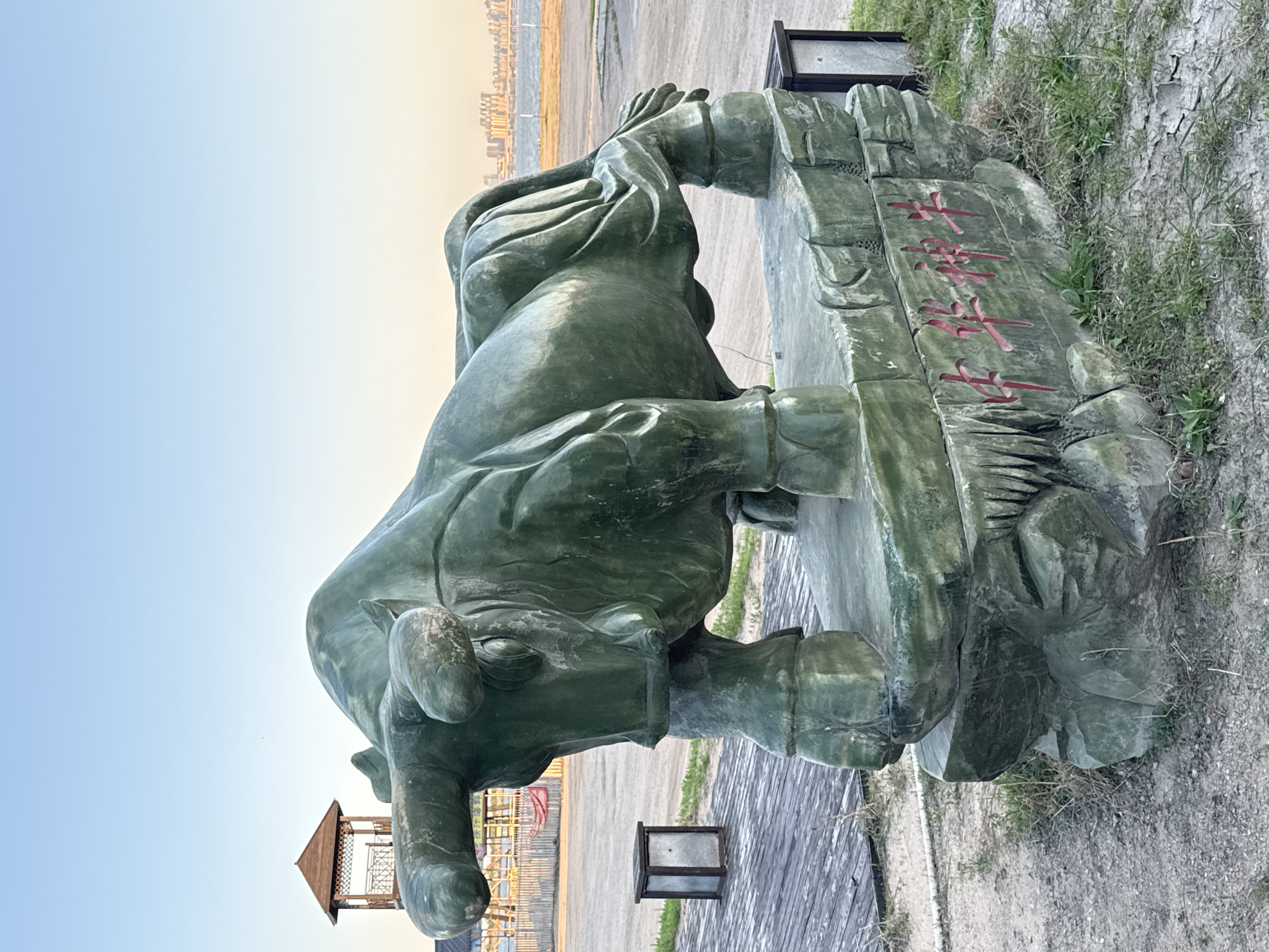 The image depicts a green, stone statue of an ox positioned sideways in an outdoor setting. The statue is on a stone base with Chinese characters inscribed in red. In the background, there are buildings and a watch tower. The ground is a mix of gravel, wooden decking, and grass. The sky is blue and clear. The location appears to be a park in the Taihe District, China.