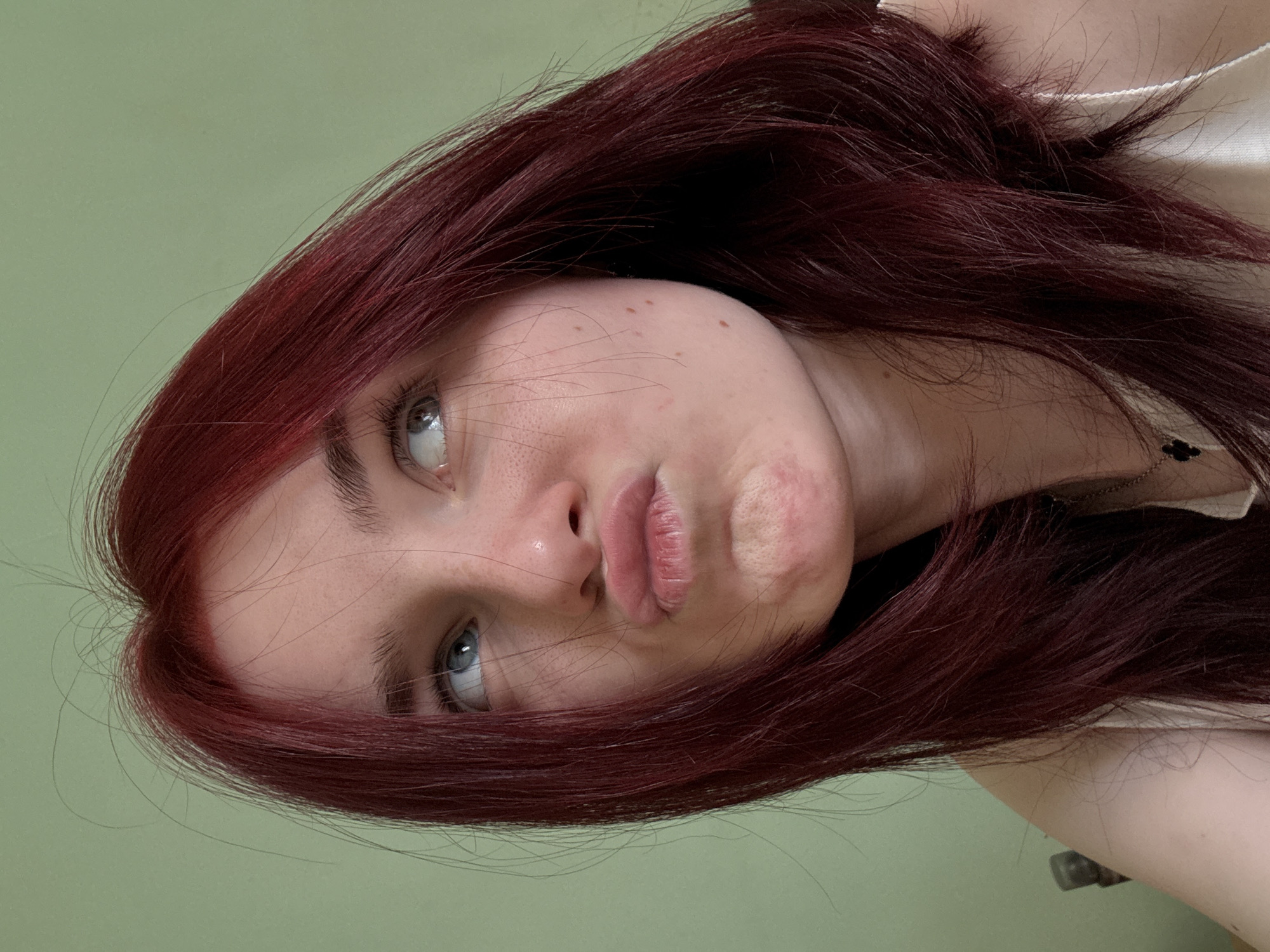 A close-up, medium shot captures a young person with reddish-brown hair pulled back and to the side, looking up and to their right with a pouty expression. Their face is turned slightly to the left, with their eyes looking upwards and to the right. Small freckles are visible on their nose and cheeks. The background is a plain, light green wall. The person is wearing a light-colored top.