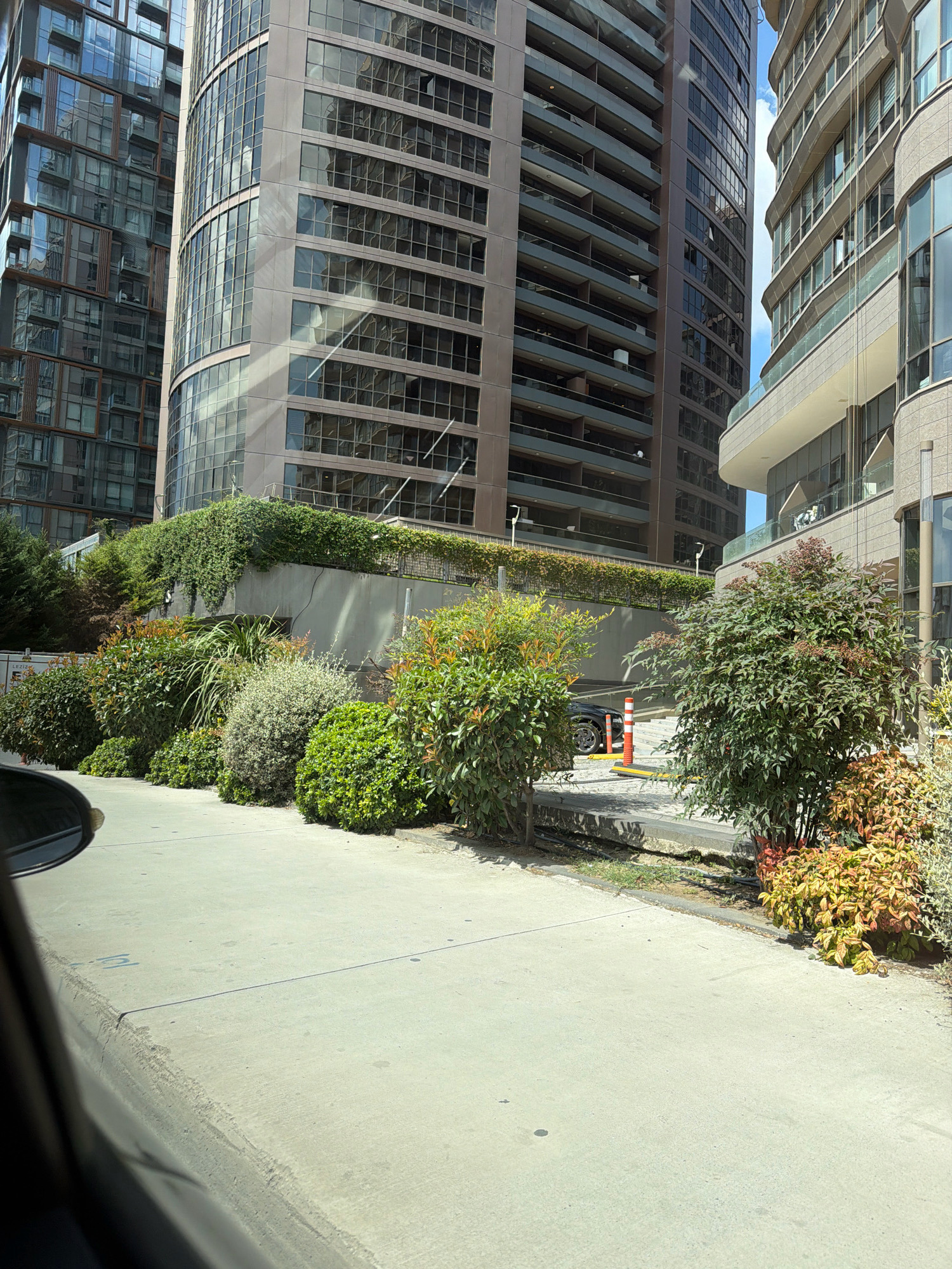 This is an eye-level, exterior, medium shot of a street scene in Bakırköy, Turkey, dominated by modern high-rise buildings and landscaping. In the foreground, a light-colored concrete walkway runs horizontally across the frame, with a dark-colored vehicle's side mirror visible in the lower left corner. Adjacent to the walkway is a manicured hedge and several bushes, some with green leaves and others displaying autumnal shades of red and yellow.
Behind the bushes, a grey concrete wall topped with a dense layer of green ivy forms a boundary. Further back, a section of a modern building with a curved facade and multiple levels of windows is visible. The building's exterior is predominantly grey and beige, with reflective windows. In the background, towering skyscrapers with numerous glass-paneled floors and balconies dominate the skyline. The buildings exhibit a mix of architectural styles, with some featuring linear designs and others having a more organic, curved appearance. The sky above is bright blue with scattered white clouds, indicating a clear day. The scene appears to be in a commercial or residential district, likely a landscaped area around office buildings or apartment complexes.