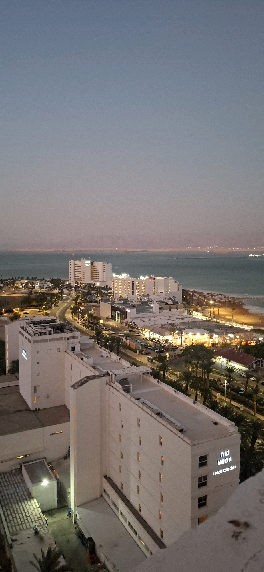 The image is an aerial, high-angle view of a coastal city at dusk. In the foreground, on the right, is a portion of a white building with illuminated windows. To the left and further back are more white buildings, one with the illuminated text "NOGA Israel Collection" on its facade. Beyond these buildings, a street with cars and palm trees is visible, leading towards a beach with umbrellas and lounge chairs. The sea is in the middle ground, with distant landmass visible on the horizon. The sky is a gradient of pale blue to light pink, indicating the time of day is either sunrise or sunset. The overall scene depicts a resort town. The city is located in the Tamar Regional Council, Israel.