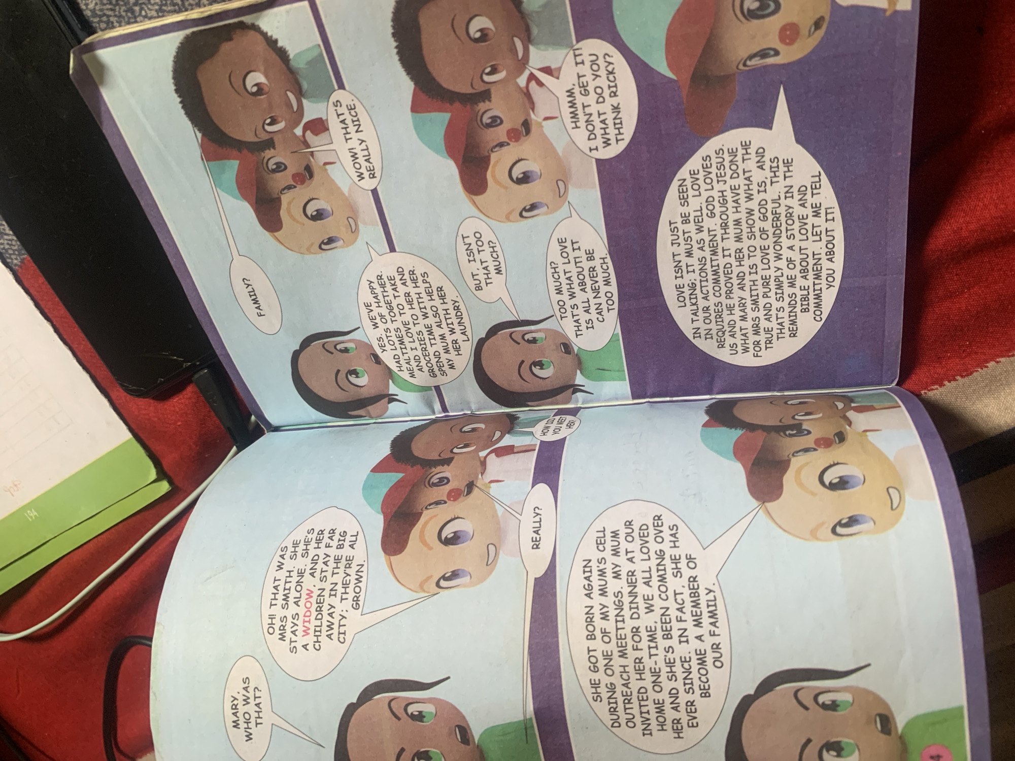 This is a photograph of an open comic book, likely a children's book. The left page depicts a comic panel with text bubbles. One character asks "MARY, WHO WAS THAT?". Another character replies "OH! THAT WAS MRS SMITH. SHE STAYS ALONE. SHE'S A WIDOW. AND HER CHILDREN STAY FAR AWAY IN THE BIG CITY; THEY'RE ALL GROWN." The right page shows a comic panel with more characters and text bubbles. One character asks "FAMILY?". Another replies "WOW! THAT'S REALLY NICE." Another character asks "BUT, ISN'T THAT TOO MUCH?". A fourth character responds "TOO MUCH? THAT'S WHAT LOVE IS ALL ABOUT! IT CAN NEVER BE TOO MUCH." A fifth character asks "HMMM, I DON'T GET IT! WHAT DO YOU THINK RICKY?". A large text bubble at the bottom of the right page reads "LOVE ISN'T JUST IN TALKING; IT MUST BE SEEN IN OUR ACTIONS AS WELL. LOVE REQUIRES COMMITMENT. GOD LOVES US AND HE PROVED IT THROUGH JESUS. WHAT MARY AND HER MUM HAVE DONE FOR MRS SMITH IS TO SHOW WHAT THE TRUE AND PURE LOVE OF GOD IS, AND THAT'S SIMPLY WONDERFUL. THIS REMINDS ME OF A STORY IN THE BIBLE ABOUT LOVE AND COMMITMENT. LET ME TELL YOU ABOUT IT!". The comic features stylized, cartoonish characters with large heads and eyes. The background is a light blue. The book is placed on a red surface, possibly a blanket or fabric, with a portion of a green object visible in the upper background. There is no indication of Jauro Yinu, Nigeria, in the image.