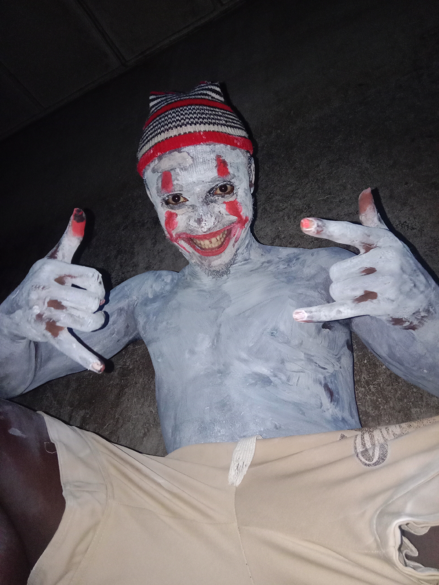 The image shows a person with painted white skin and a red clown smile, wearing a red and black striped beanie. The person is lying down with arms raised, hands making a "rock on" gesture. The fingernails are painted red. The person is wearing light-colored shorts. The background is dark and appears to be a concrete surface. The location is Unknown, Unknown.