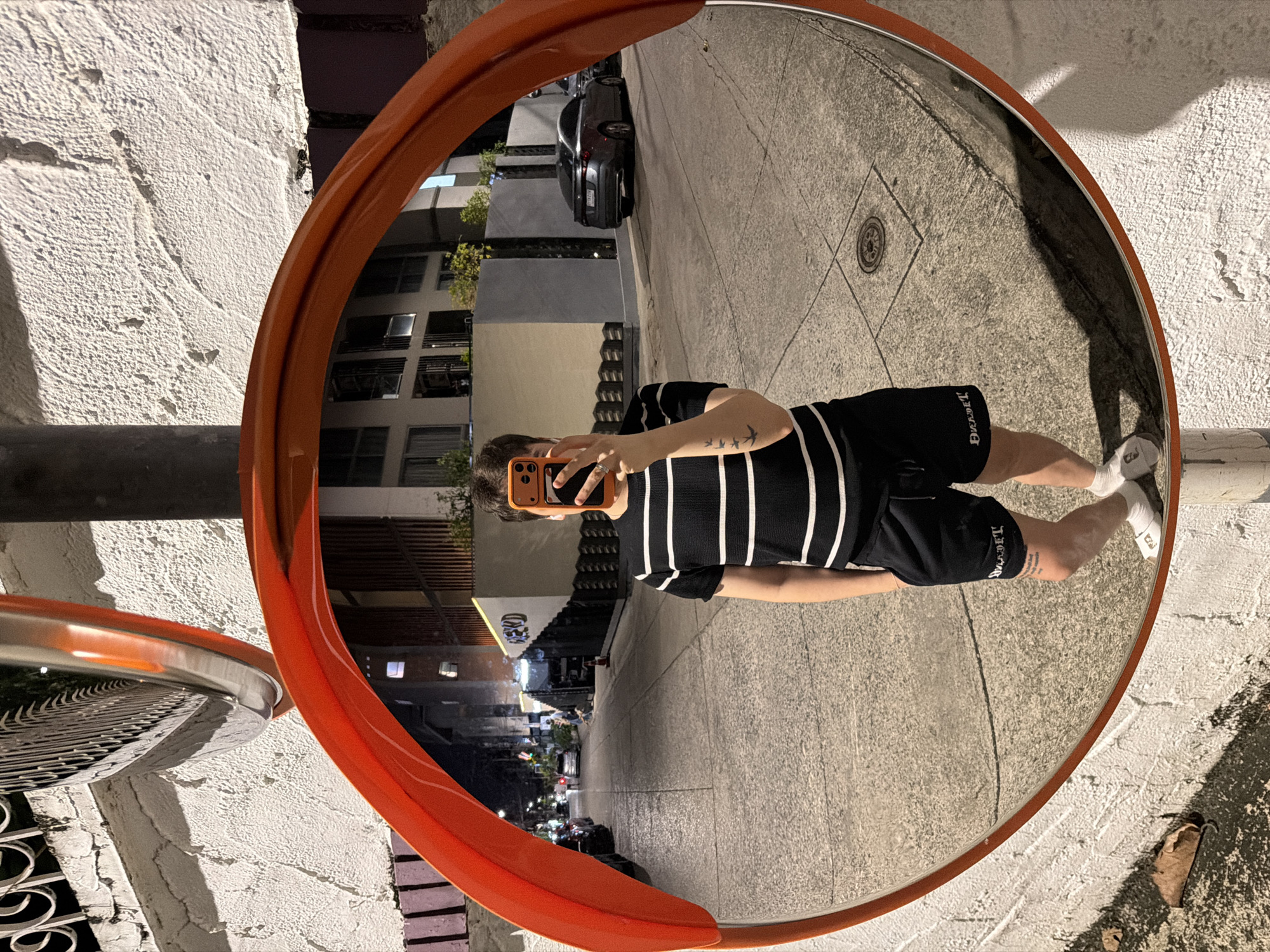 The vertical image centers on a large, orange-framed convex safety mirror reflecting a person taking a selfie. The person, oriented horizontally within the image frame, holds a smartphone with an orange case, obscured by their hand, up to their face. They wear a black and white striped short-sleeved top, black shorts with stylized text, possibly "DUKDET," on the left leg, white socks, and white athletic shoes. A tattoo of three small birds in flight is visible on their left forearm.
The mirror's wide-angle reflection depicts an urban street scene at night or dusk in Bangkok, Thailand, illuminated by artificial lighting. A dark-colored car is parked on the street, and multi-story buildings with windows and balconies line the background, interspersed with some trees. The ground is paved with concrete slabs, and a circular manhole cover is visible.
Outside the mirror, the immediate foreground and edges of the image feature a rough-textured, light-colored wall. The scene captures a casual moment of self-reflection in a bustling city environment, highlighted by the distinctive distortion of the convex mirror.