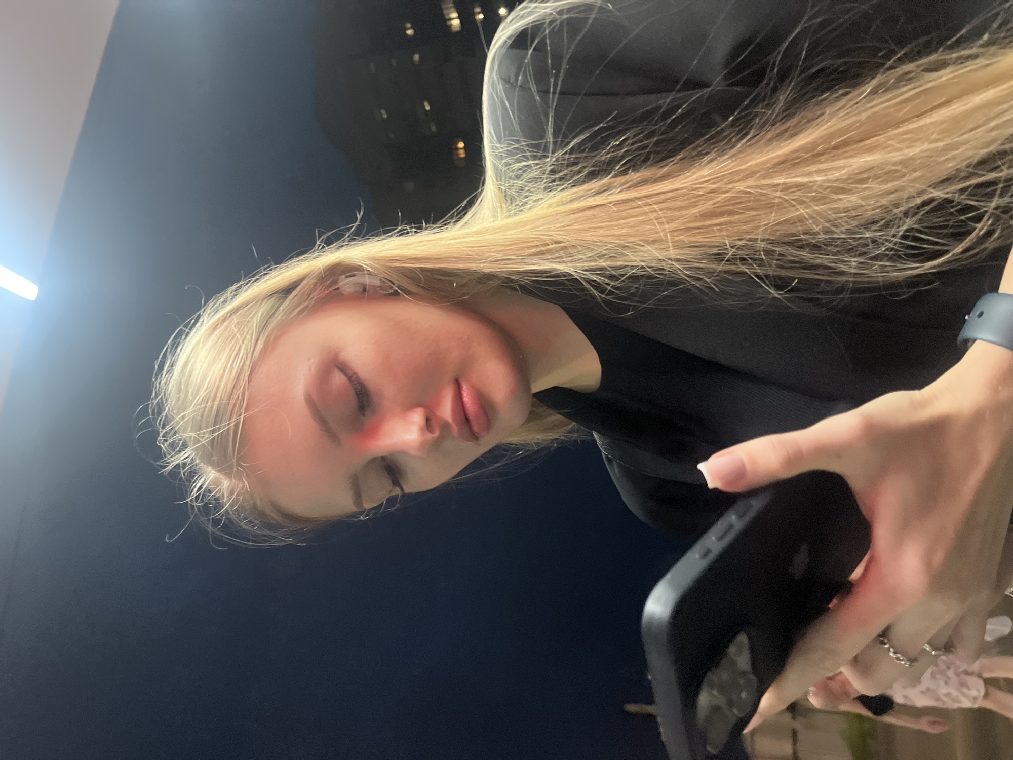 An outdoor nighttime medium shot captures a young Caucasian woman with long blonde hair, wearing a black blazer over a black top and white earbuds. She is looking down at a black smartphone held in her right hand. Her left hand, adorned with rings and manicured nails, is also visible near the phone. A red light illuminates her cheek and nose. The background is dark, with a hint of a building with lit windows in the distance. The scene appears to be in a city environment. There is no specific indication of the location being Perm, Russia.