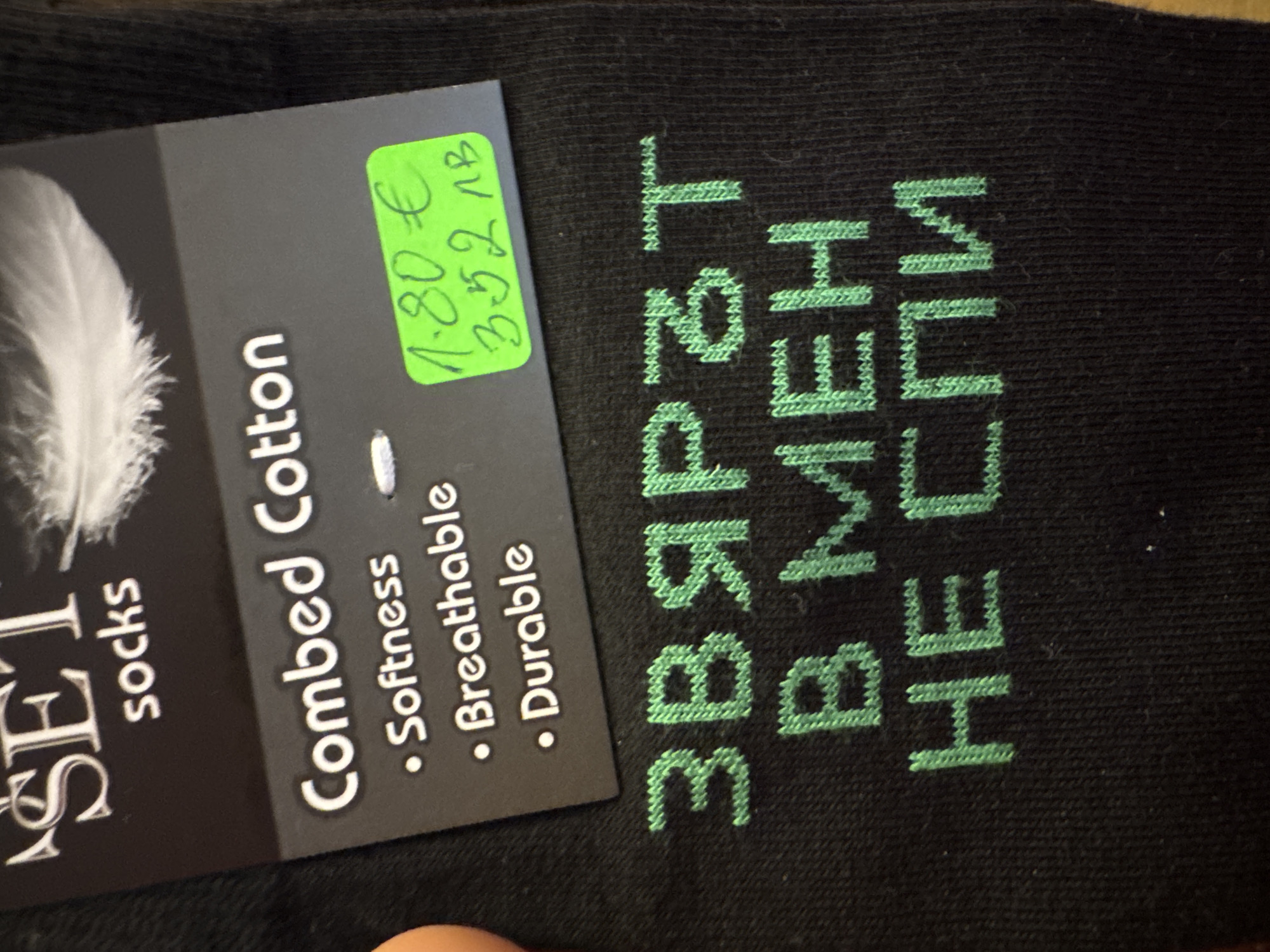 The image shows a close-up of a pair of black socks with a tag attached. The tag identifies the brand as "SET socks" and describes the material as "Combed Cotton," highlighting its softness, breathability, and durability. A green price tag sticker displays "1.80€" and "3.52 MB." Embroidered on the socks in green Cyrillic script is the phrase "ЗВЯРЪТ В МЕН НЕ СПИ," which translates to "The beast in me does not sleep." The setting appears to be indoors, possibly in a store or at home. No specific information indicates the location of Burgas, Bulgaria, but the use of Cyrillic script suggests the socks may be sold in a Bulgarian-speaking region.