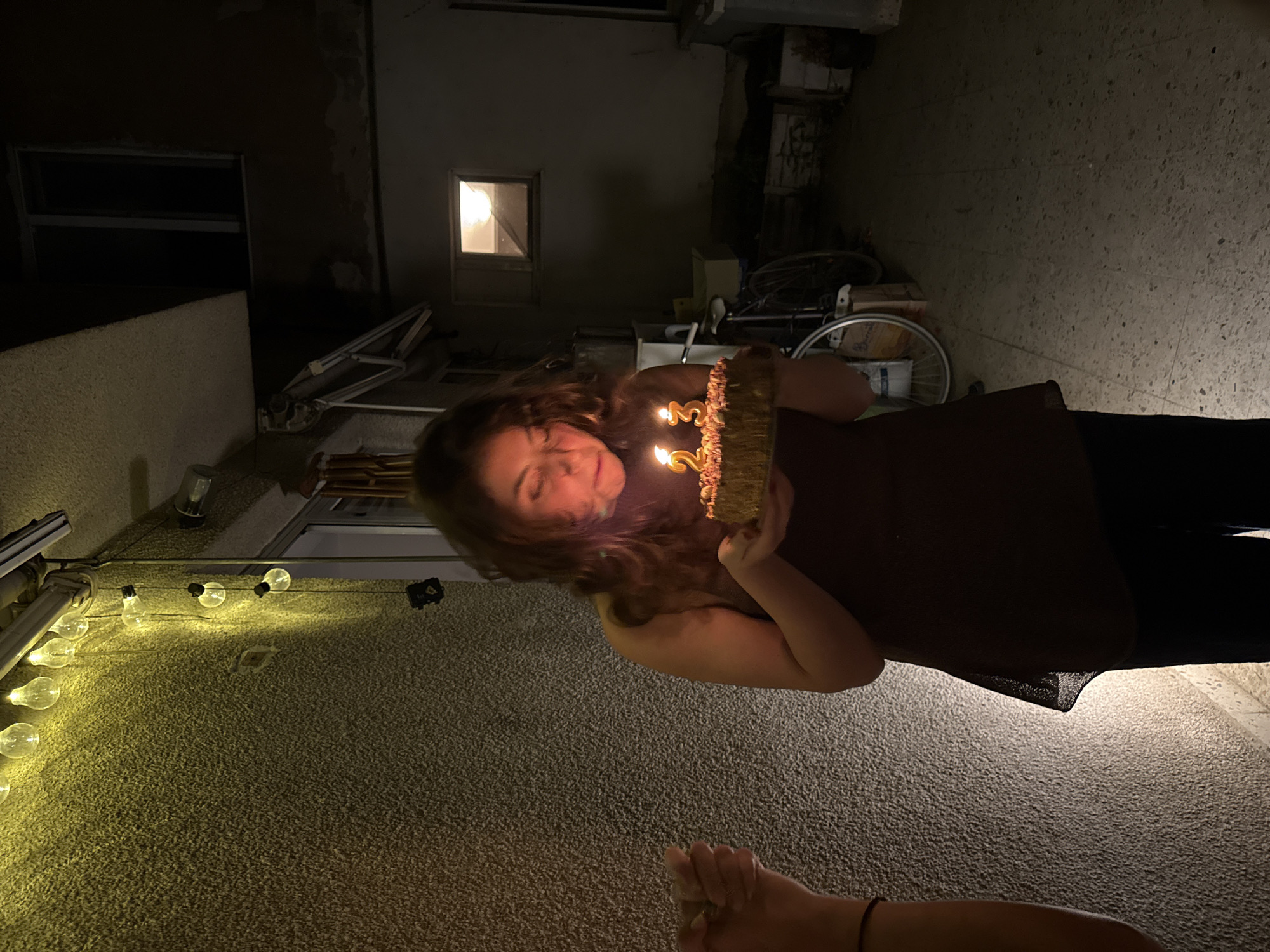 A woman holds a birthday cake with candles that read '23' outdoors at night, possibly in Sofia, Bulgaria, based on architectural styles. She is wearing a dark top and dark pants. The background shows a building with a window and string lights overhead. A bicycle is partially visible. Another person's foot and ankle are visible in the lower foreground.