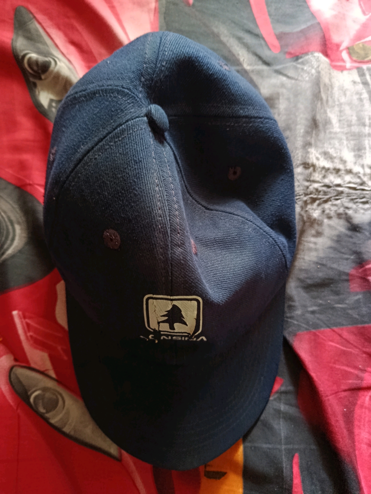 A dark blue baseball cap with a white and black logo featuring a silhouette of a tree is placed on a red and black patterned fabric. The logo is on the front of the cap. The cap is positioned at an angle, with the brim facing downwards and to the right. The fabric appears to be soft and possibly a blanket or bedspread, with a vibrant red section and a darker, more subdued patterned section. The lighting suggests an indoor setting, and the overall impression is of a casual placement of the cap. The image does not provide any information to determine its location in Jambearum, Indonesia.