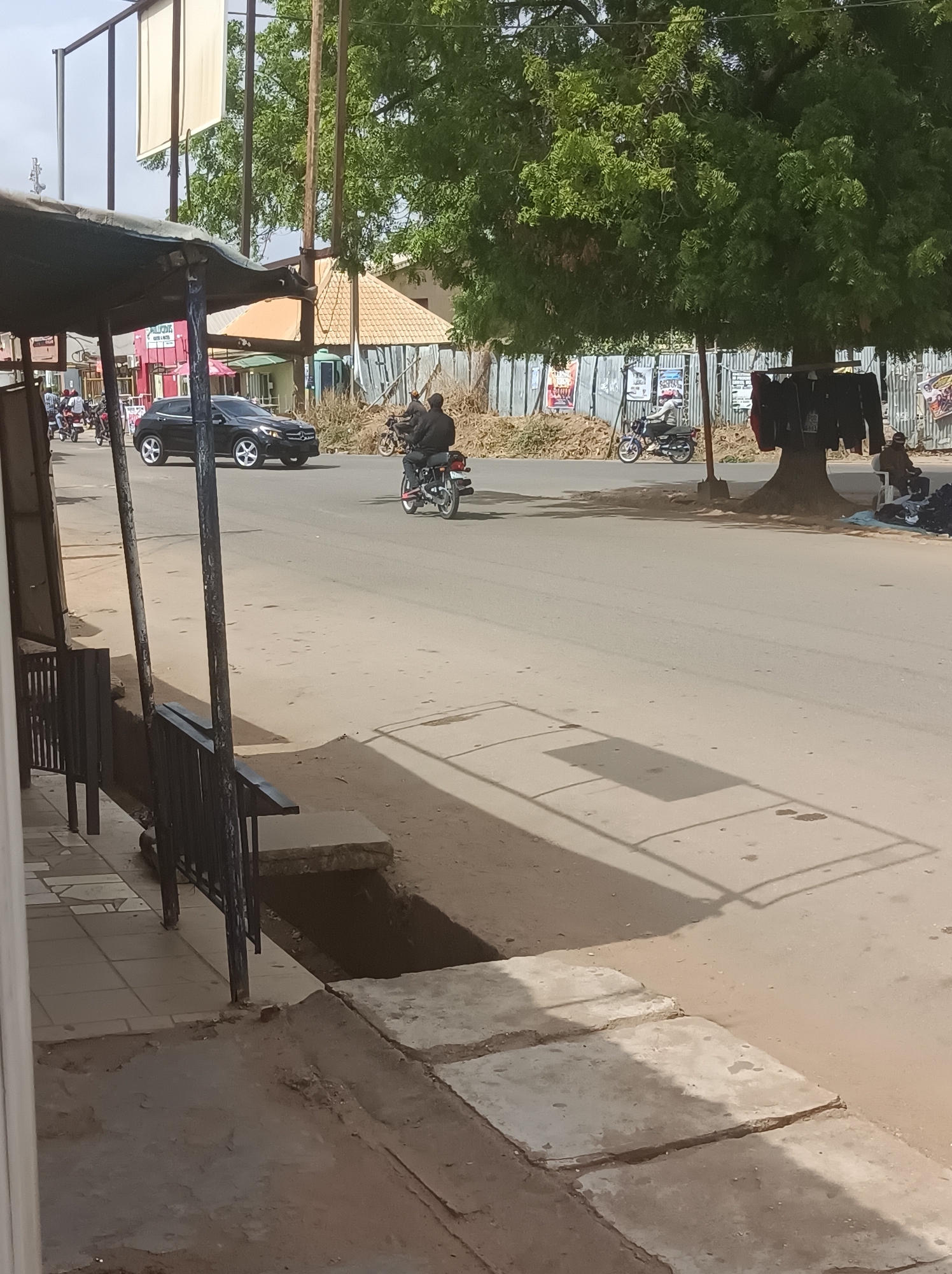 This image depicts a bustling street scene in Narayi, Nigeria, under bright, sunny conditions, likely mid-morning or early afternoon as indicated by the long shadows stretching across the road and foreground.
In the foreground on the left, a portion of a rustic, metal-roofed shelter extends into the frame. The main subject is a paved road where a black Mercedes-Benz SUV is driving left-to-right. Several motorcycles are also present; one motorcyclist in a black jacket is in the mid-ground, and another in a lighter top is further down the road on the right.
On the right side of the road, a large, leafy tree provides shade. Clothes, including jackets and shirts, are hung from its branches, suggesting a roadside vendor's setup. A person is seated on a low stool beneath this tree, presumably attending to the merchandise. Behind the tree, a corrugated metal fence and piles of brush line the street.
The immediate foreground on the left features a concrete sidewalk section with a metal railing, leading to an open drainage ditch or gutter. In the background, buildings with distinct tiled roofs and other structures are visible, and large, blank billboards stand against the bright sky. The scene captures a slice of daily life, combining transportation with informal commerce. No legible text is visible on any signs or posters.