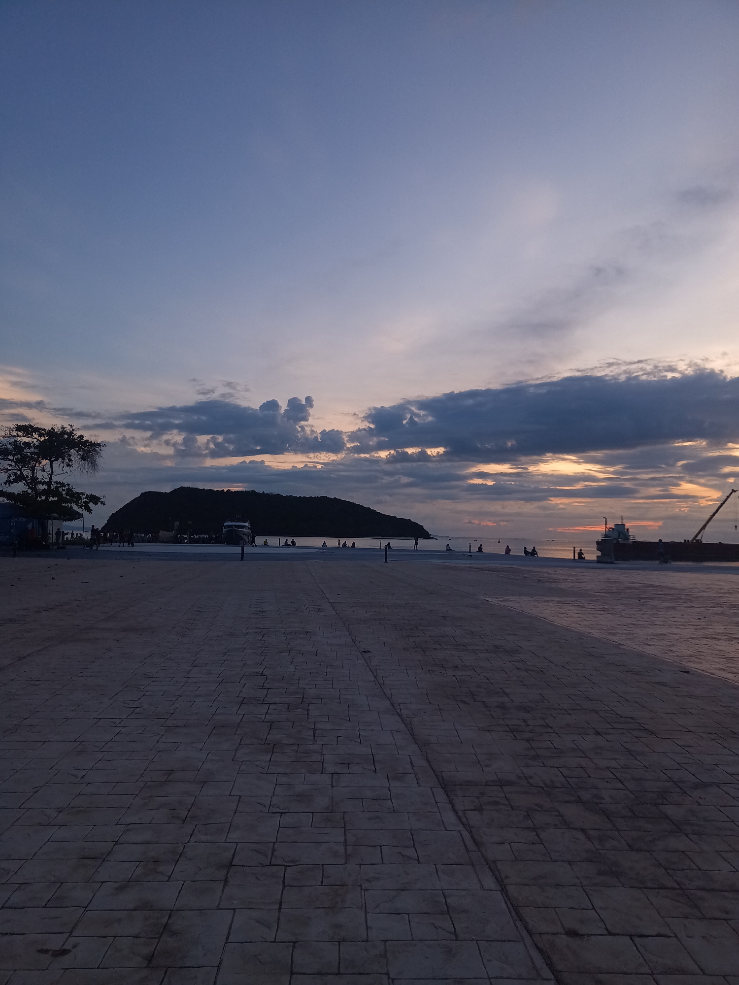 The image depicts a coastal scene at dusk in the area of Koh Phangan, Thailand. A paved promenade runs from the foreground into the middle ground, with large, rectangular stone tiles forming the surface. On the left side of the promenade, a silhouetted tree stands near a building. Further into the middle ground, a group of people are scattered along a raised walkway or pier. A dark, island-like landmass is visible in the distance, and a boat is docked near it. To the right, a large cargo ship with a crane is positioned in the water. The sky above is a gradient of blue and purple hues, with scattered clouds that are illuminated by the setting sun, creating warm orange and pink tones. Several small figures, presumed to be people, are visible sitting or standing on the walkway and the shoreline.